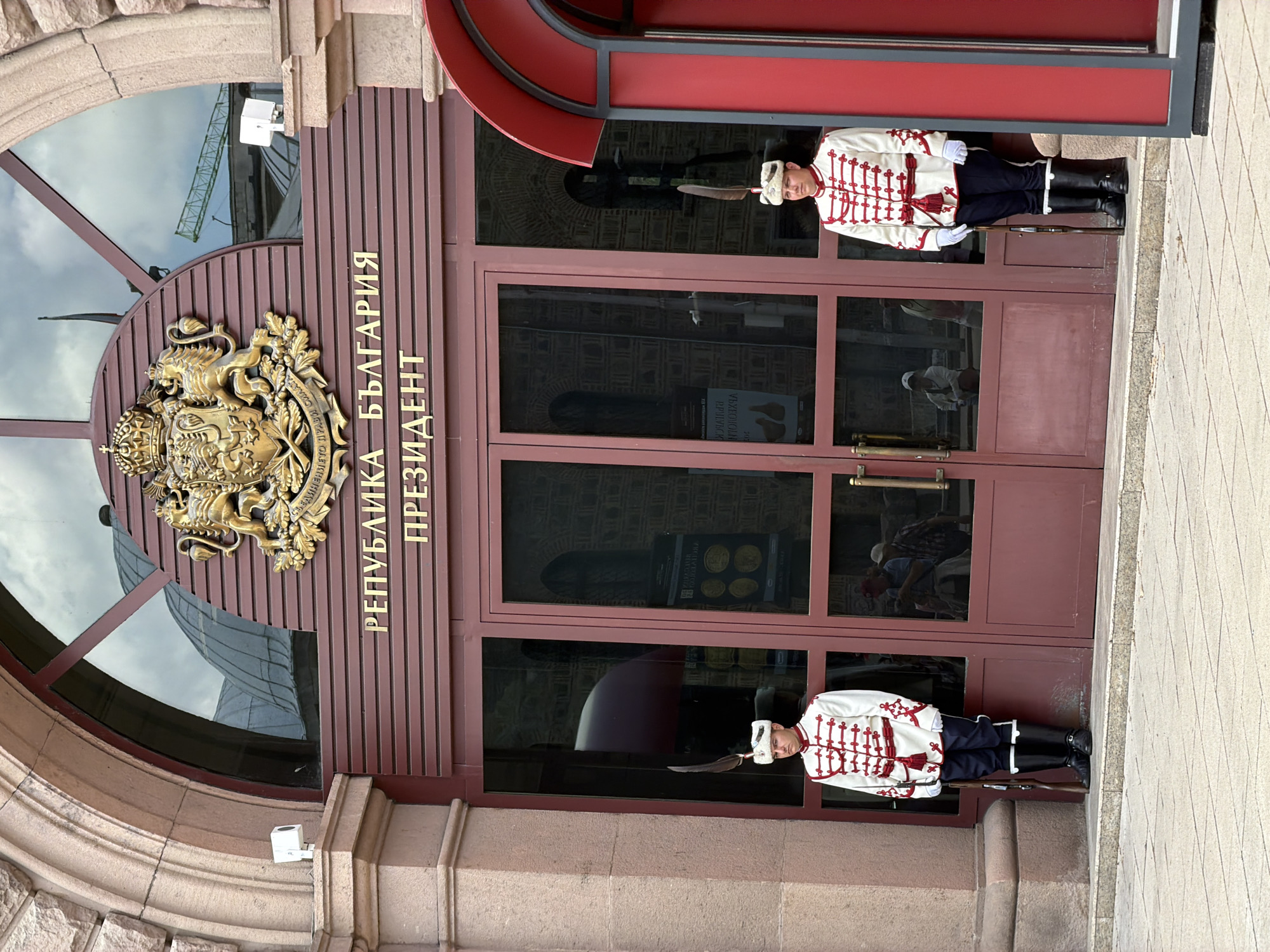 A daytime eye-level shot captures two ceremonial guards standing sentry outside a grand building entrance in Sofia, Bulgaria. The building features an ornate stone archway and dark wooden or metal double doors. Above the doors, prominent golden Cyrillic text reads "РЕПУБЛИКА БЪЛГАРИЯ ПРЕЗИДЕНТ," translating to "REPUBLIC OF BULGARIA PRESIDENT."
To the left of the doors, within a large arched window, a golden, ornate Bulgarian coat of arms is displayed, depicting two rampant lions supporting a shield with a third lion, all beneath a crown. The glass surfaces of the doors and window reflect the bright, slightly overcast sky and a construction crane, indicating an outdoor urban setting. A white security camera is mounted on the stone archway to the left.
The two male guards are positioned symmetrically on either side of the entrance, standing at attention. They are dressed in distinctive ceremonial uniforms consisting of white tunics with intricate red braiding and epaulets, dark trousers, polished black tall boots, and white hats adorned with feathers. Each guard holds a long rifle or musket vertically at their side, maintaining a rigid and formal posture. The scene conveys a sense of official duty and national presence.