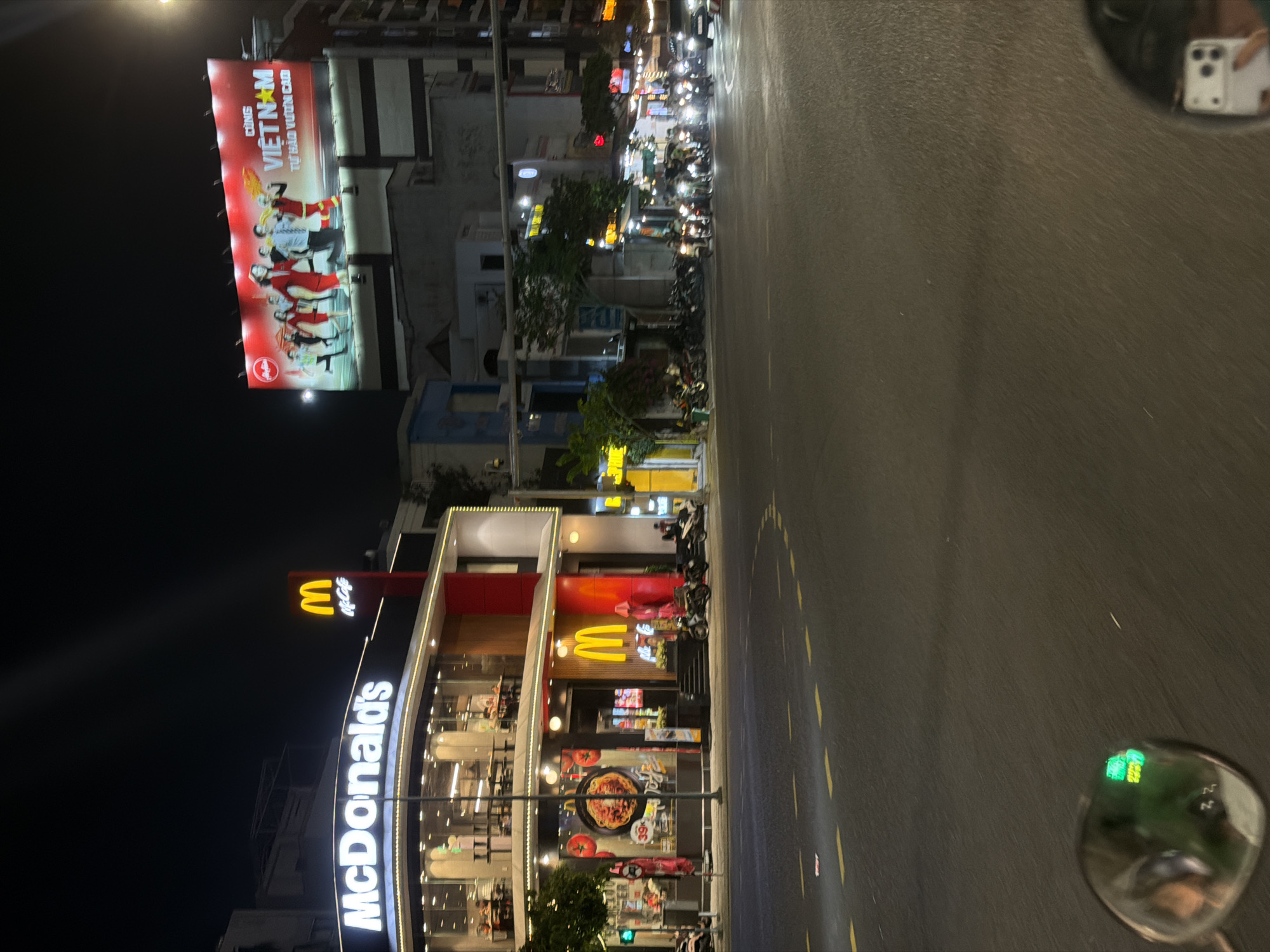 The image captures a nighttime street scene in Đà Nẵng, Vietnam. Dominating the left side of the frame is a brightly lit McDonald's restaurant with its iconic signage and large glass windows, showcasing patrons inside. To the right, a large billboard displays an advertisement for "Vietnam" with people in celebratory poses and text in Vietnamese. The street is paved and marked with yellow lines, indicating a road. Several motorcycles are parked and in motion, suggesting typical urban traffic. The overall environment appears to be an active commercial area at night, illuminated by streetlights, building lights, and the advertisement's glow. The perspective suggests the photo was taken from a moving vehicle, possibly a motorcycle, given the presence of a rearview mirror in the foreground reflecting a digital display and a cell phone.