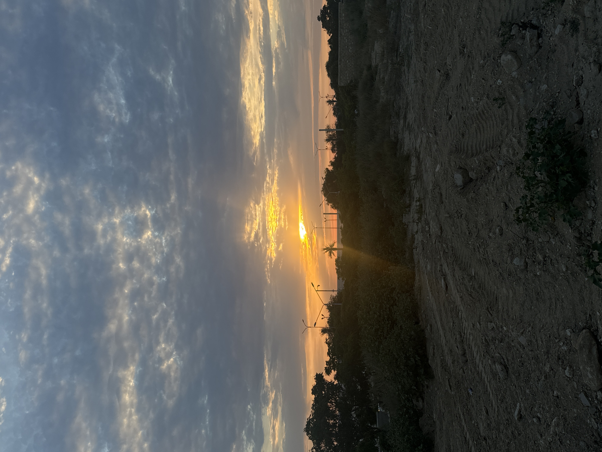 The image depicts an outdoor scene, likely at dusk or dawn, overlooking a somewhat desolate area with sparse vegetation and rough, unpaved ground in the foreground. In the midground, a line of trees and bushes forms a natural border, with several streetlights interspersed among them. A hint of water or distant land can be seen beyond the foliage. The sky is filled with dramatic clouds, tinged with the warm hues of the setting or rising sun, which peeks through the cloud cover, casting visible rays of light. The overall impression is one of an undeveloped or transitional space on the outskirts of a city, possibly near a coast given the light colors on the horizon. The lack of specific landmarks makes pinpointing Aktau, Kazakhstan, impossible based on visual evidence alone.