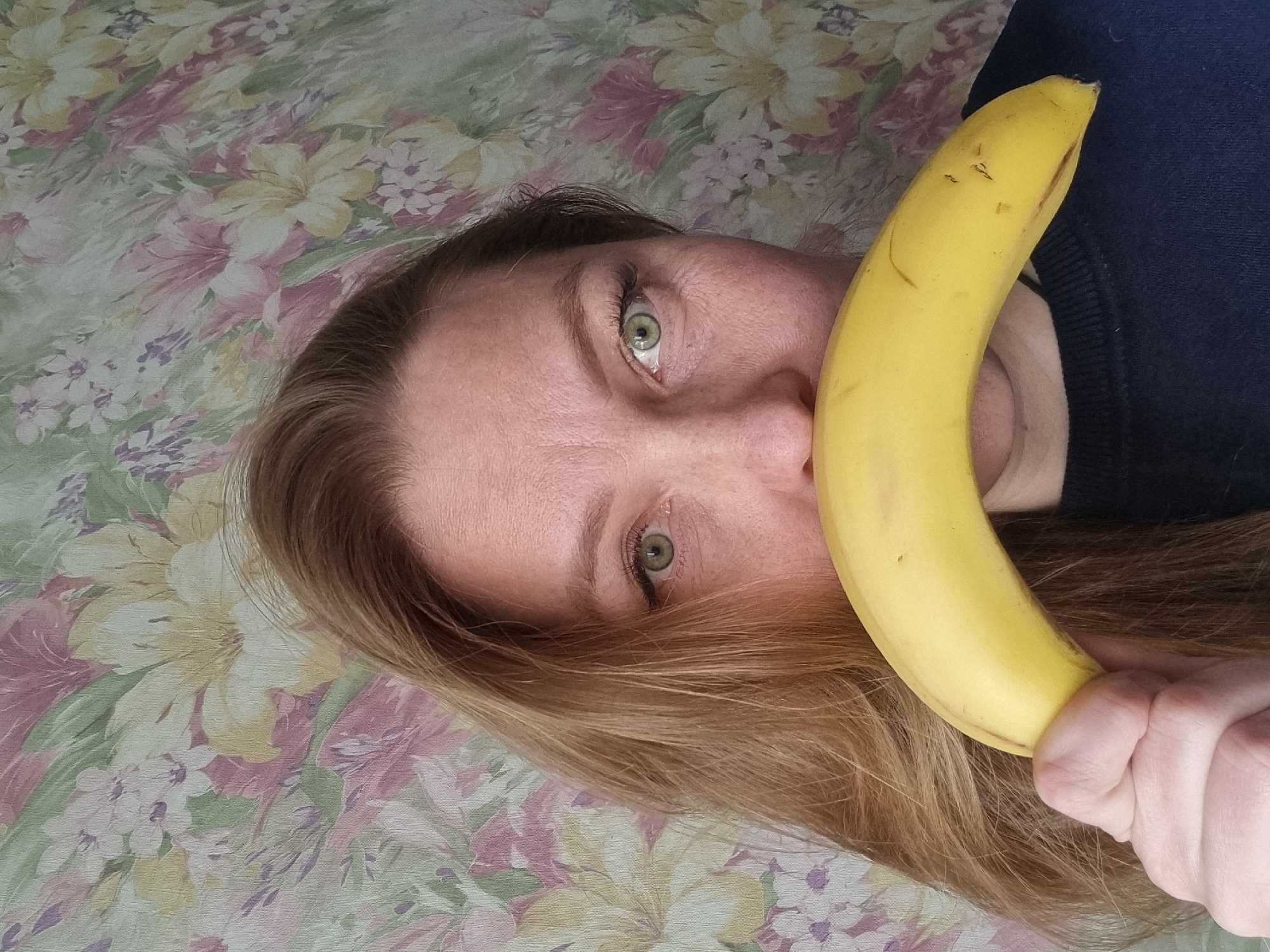 A female individual, appearing to be an adult of Caucasian ethnicity, is depicted in an indoor setting in Chaikovsky, Russia. She possesses light brown hair and green eyes, both directed towards the viewer. Her lower facial features, including the nose and mouth, are obscured by a horizontally positioned, ripe yellow banana. The individual's left hand is visible, gripping one end of the banana. She is wearing a dark blue crew-neck garment. The background consists of wallpaper featuring a floral pattern with prominent light green, yellow, pink, and purple elements.