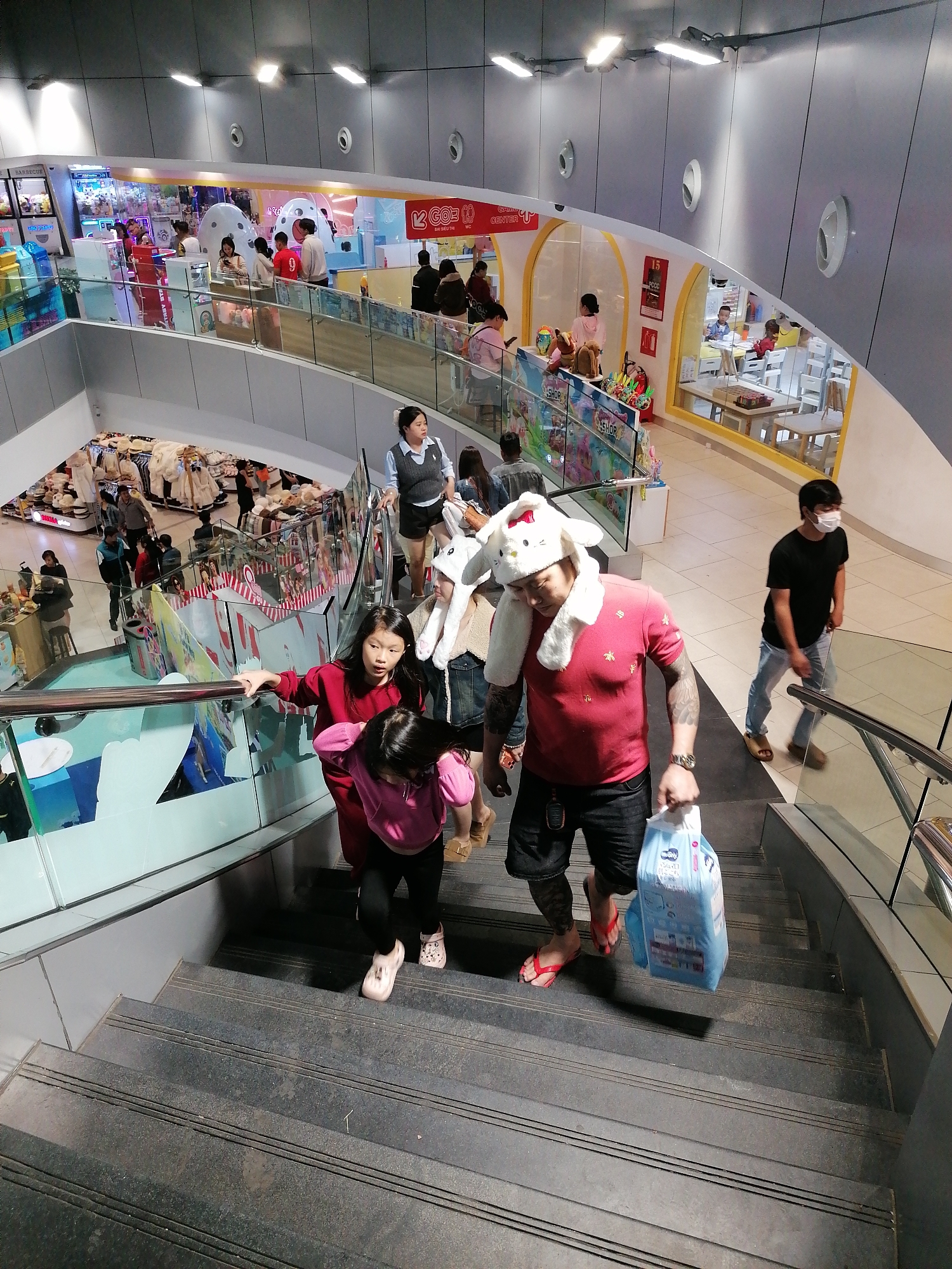 The image captures a scene in a shopping mall, possibly in Da Lat, Vietnam. A man in a red t-shirt, black shorts, and red flip-flops, wearing a novelty white bunny hat, is descending a set of gray stairs. He carries a large package of baby diapers in his right hand. Next to him is a woman in a red sweater dress and a small child in a pink top and black pants with white clogs. The woman is holding the girl's hand. The man has tattoos on his right arm. A woman in a vest and shorts, along with others, are visible above them on an upper level. Another man wearing a black t-shirt, light blue jeans, and a face mask is walking on the upper level. Various shops and a play area are visible in the background, suggesting a family-oriented environment. The architecture features curved lines and metallic accents, with bright, even lighting.