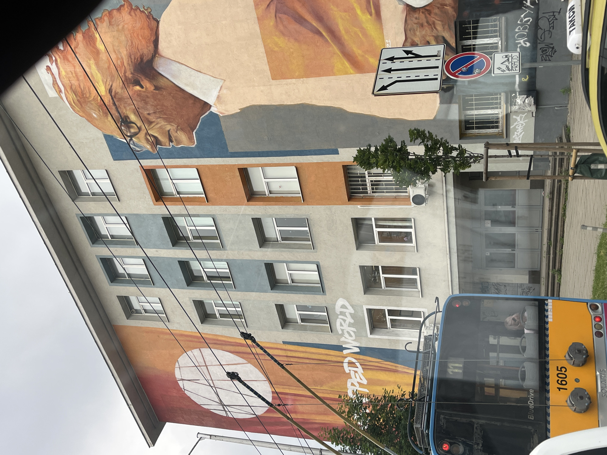 A mural of an elderly man with fiery orange hair, wearing glasses and a light-colored collared shirt, is painted on the side of a multi-story building in an urban setting. The mural occupies a significant portion of the building's facade, which is painted in sections of grey, blue, and orange. In the foreground, the rear of a blue and yellow trolleybus is visible, displaying the number 11 and "BlueDrive." The trolleybus has a license plate reading "1605" and "SOLARIS." Street signs, including one indicating multiple lanes with arrows and a no parking sign, are mounted on the building. Graffiti is present on the lower section of the building and on a white box attached to it. The sky is overcast. A young tree is planted in front of the building.