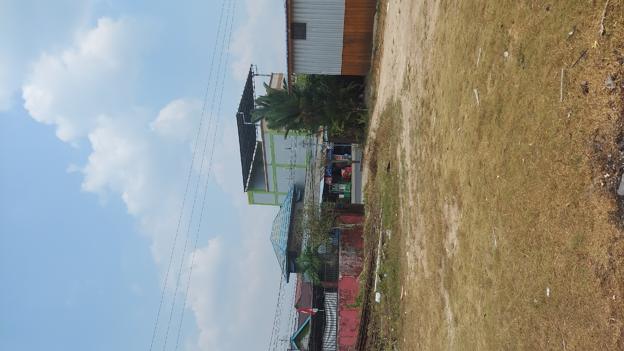 The image depicts a street scene in Tanjung Redeb, Indonesia. In the foreground, dry, brown grass covers the ground, with scattered debris. A dirt road curves into the middle ground, leading towards a collection of buildings. To the left, a building with a blue tiled roof and a red-painted wall is visible, with a metal fence in front of it. A small tree and some plants grow near this building. Further back, a multi-story building with green and white sections and a dark roof structure stands prominently. To the right, a long, corrugated metal building with orange and silver panels is present. The background shows a clear blue sky with scattered white clouds and overhead power lines. The scene appears to be a commercial or residential area.