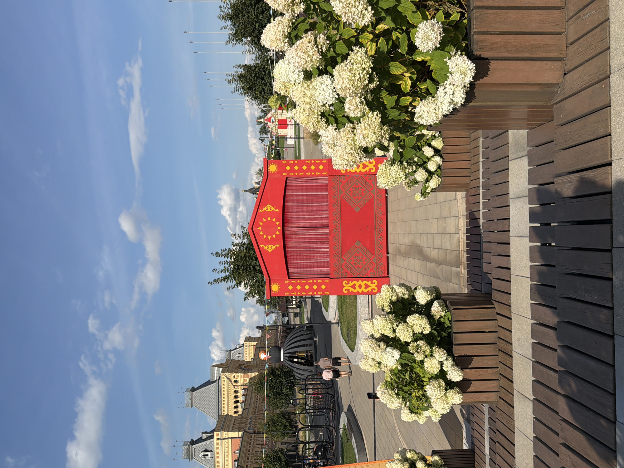 The image shows a bright, sunny day with a clear blue sky and scattered white clouds. In the foreground, two large wooden planters overflowing with white hydrangeas are placed on a wooden bench. Behind the planters, a vibrant red, open-air stage with yellow decorative elements and red curtains stands on a paved walkway. Two people, a man and a woman, walk away from the camera on the walkway towards a large, ornate building with a yellow facade and grey, pointed roofs in the background. To the left of the building, a large, dark, dome-shaped structure is visible. The scene is likely located in the city of Nizhny Novgorod, Russia, possibly at a public square or event venue, given the architecture and the temporary stage setup.