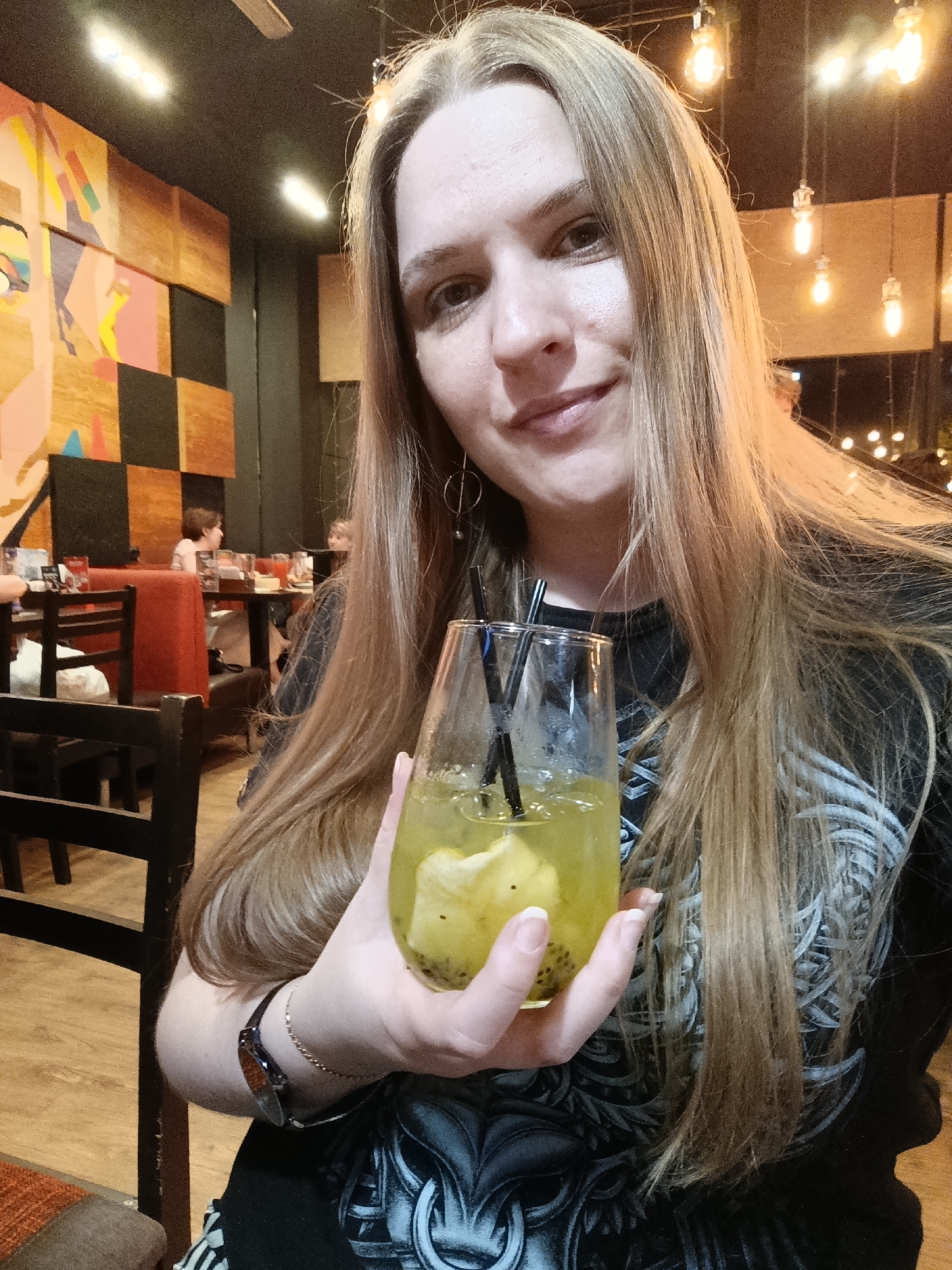 A woman is holding a glass of kiwi lemonade in a restaurant in the city of Unknown, Unknown. The glass has a wide bottom and tapers towards the top. Inside, there are slices of kiwi, ice, and two black straws. The liquid is a pale green color. She is fair-skinned with long blonde hair that falls past her shoulders. She is wearing a black t-shirt with a white design. On her left wrist, she has a watch with a brown band and a thin gold bracelet. She is smiling slightly and looking at the camera.
The background shows a restaurant setting. There are people sitting at tables behind her. The walls have various decorations, including a large, colorful abstract painting with geometric shapes. Above the woman, there are several exposed light bulbs hanging from the ceiling. The lighting is somewhat dim, creating a warm atmosphere.