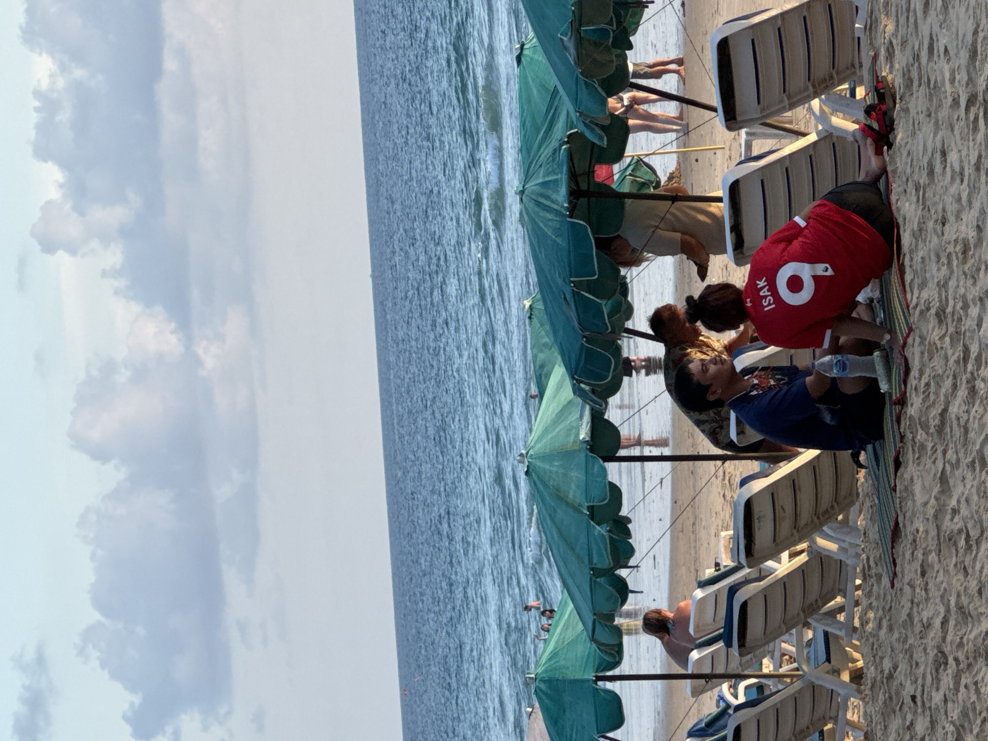 This is a wide shot of Karon Beach in Karon, Thailand, taken during daylight. The scene captures a stretch of sandy beach with several rows of empty green beach umbrellas and white lounge chairs. In the foreground, a man and a woman are sitting on a striped mat on the sand. The woman is wearing a red soccer jersey with the name "ISAK" and the number "9" on the back. A bottle of water is visible next to them. In the background, more beachgoers are visible, some in the water and some walking along the shore. The ocean is a deep blue, with gentle waves rolling onto the beach under a partially cloudy sky. The overall impression is of a relaxed beach atmosphere.