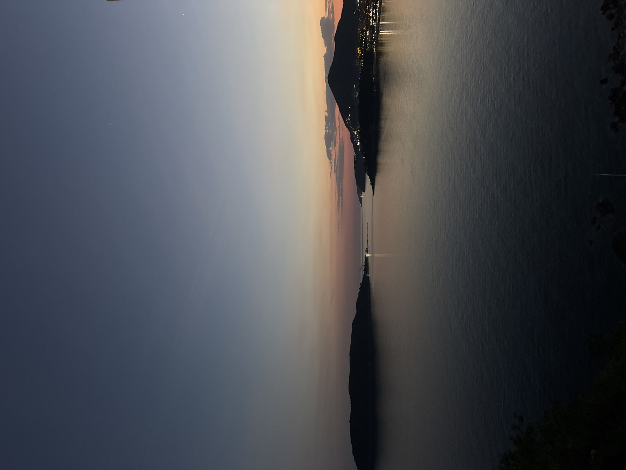 The image shows a twilight landscape of the Adriatic Sea. A body of water, presumably the Adriatic, stretches towards the horizon. Along the right side of the sea, there is a shadowy, mountainous coastline with scattered lights indicating a settlement, likely Rafailovići, Montenegro. A dark silhouette of a mountain range is prominent along the horizon, with a colorful gradient in the sky, transitioning from dark blue to pink and orange hues, indicating either sunrise or sunset. The overall scene captures a calm, serene moment in a coastal environment.