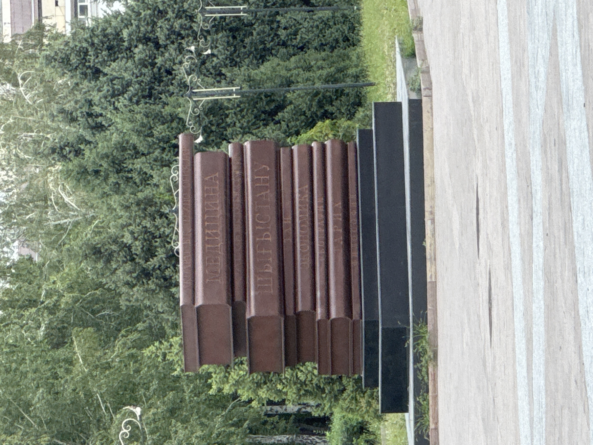 The image shows a monument resembling a stack of books in a park in Almaty, Kazakhstan. The monument is made of dark red material, with each book inscribed with text in Cyrillic. The top book reads "МЕДИЦИНА" (Medicine), the second reads "КЫРГЫЗСТАНУ" (Kyrgyzstan), and the third appears to be "ЭКОНОМИКА" (Economics). The monument stands on a black stone pedestal. In the background, lush green trees and a building are visible. The ground in front of the monument is paved, and there are white stripes indicating a pedestrian crossing. The scene is set in an outdoor, public space, likely a park or square.