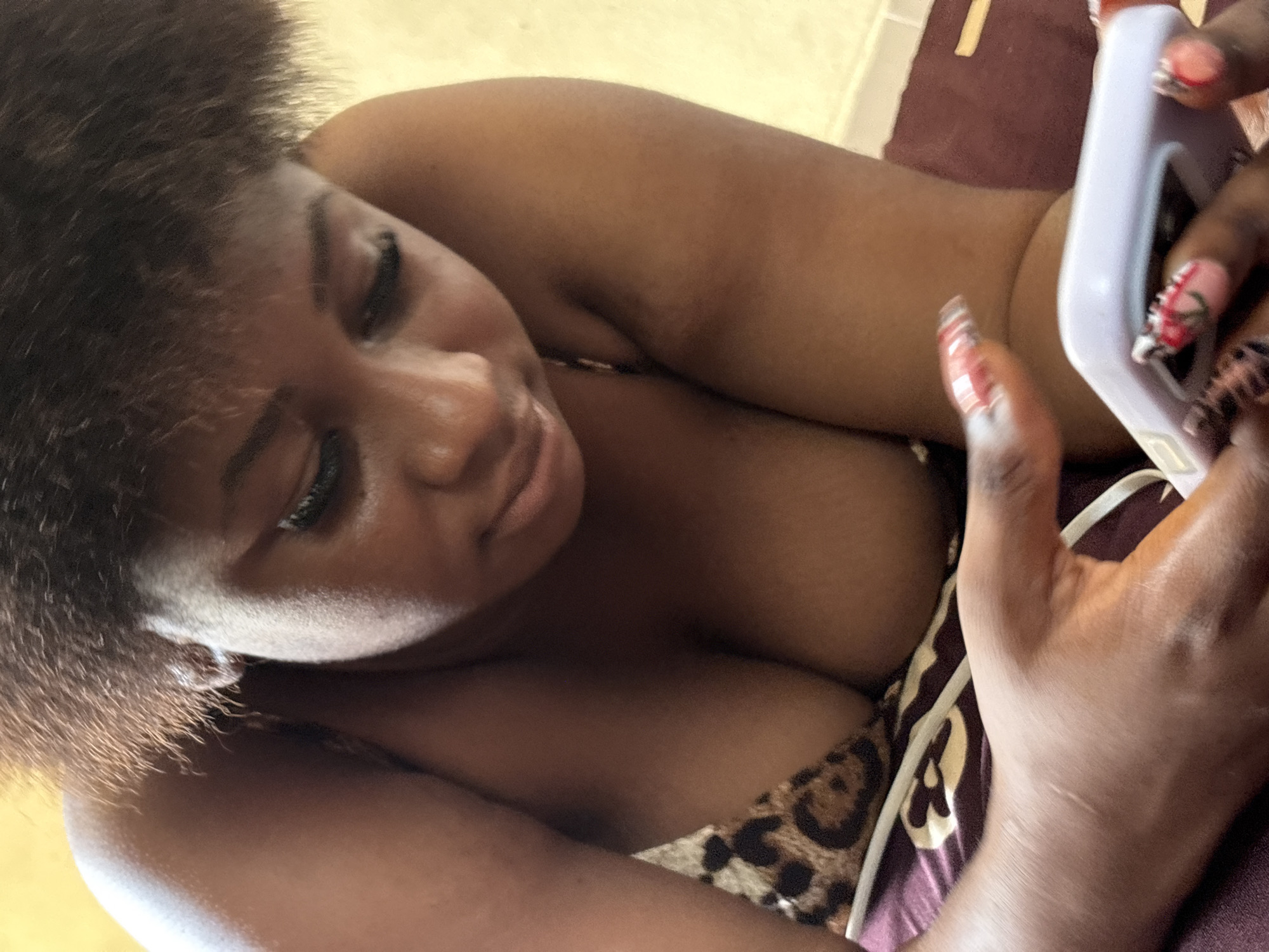 The image captures a person, identified as female, reclining indoors, engaging with a smartphone. The subject has dark skin and full, dark, curly hair that frames her face. Her head is tilted, and her gaze is directed downwards, focused on the light-colored smartphone she holds in both hands. A gentle, perhaps contemplative or engaged, expression is visible on her face.
Notable details include her long, intricately decorated acrylic nails, which feature red, white, and black patterns on an opaque base. She is wearing a leopard print garment, possibly a top or bikini, which is visible across her chest. The background suggests an indoor setting, likely a bedroom or living area, with a maroon patterned fabric, possibly a blanket or cushion, visible beneath her. The lighting indicates sufficient illumination, though the exact time of day is not discernible. The activity centers around the person's interaction with the smartphone. The location context provided for this image is Ikot Ekpene, Nigeria. There is no visible text in the image.