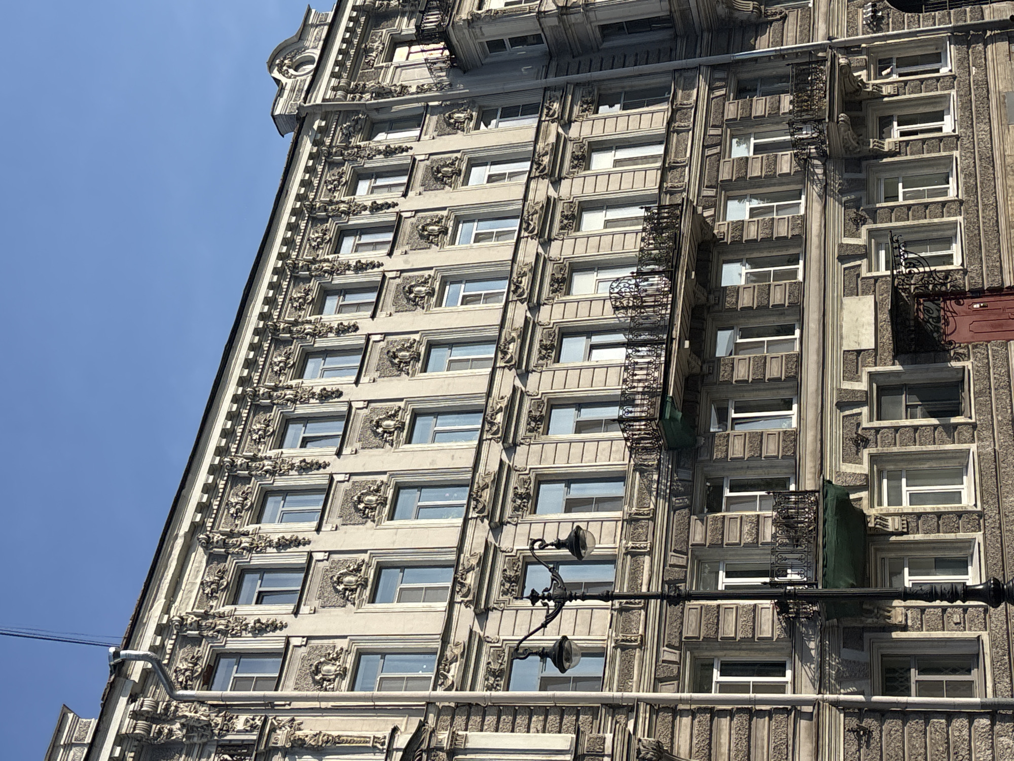 The image shows a close-up, low-angle view of an ornate building facade in Saint Petersburg, Russia, under a clear blue sky. The building exhibits a classical architectural style with numerous decorative elements. These include elaborate moldings, carved reliefs, and cornices. The facade features multiple rows of windows, many with classical surrounds and some with decorative balconies. A street lamp with two globes stands prominently in the foreground, partially obscuring some of the building's details. The stone texture of the building's lower levels suggests a rough, possibly weathered, finish. The overall impression is one of grand, historical architecture.