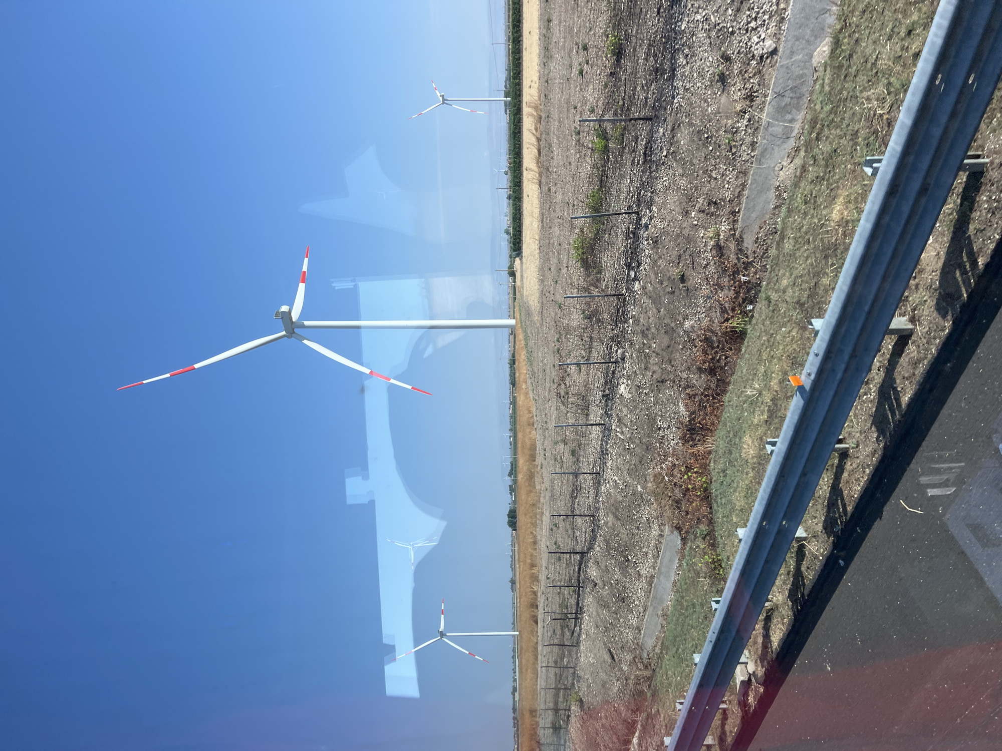 The image depicts a landscape view near Orta Nova, Italy, characterized by a clear blue sky and a vast field dotted with wind turbines. In the foreground, a metal guardrail runs horizontally across the frame, casting a distinct shadow on the asphalt road below. The field itself is a mix of dry, golden grass and patches of bare earth, divided by a wire fence with metal posts. Further in the background, more wind turbines are visible, some appearing as distant silhouettes against the horizon. The reflections on a glass surface, possibly a window, show blurred shapes of the wind turbines and the sky. The overall scene conveys a rural, open environment with a focus on renewable energy infrastructure.