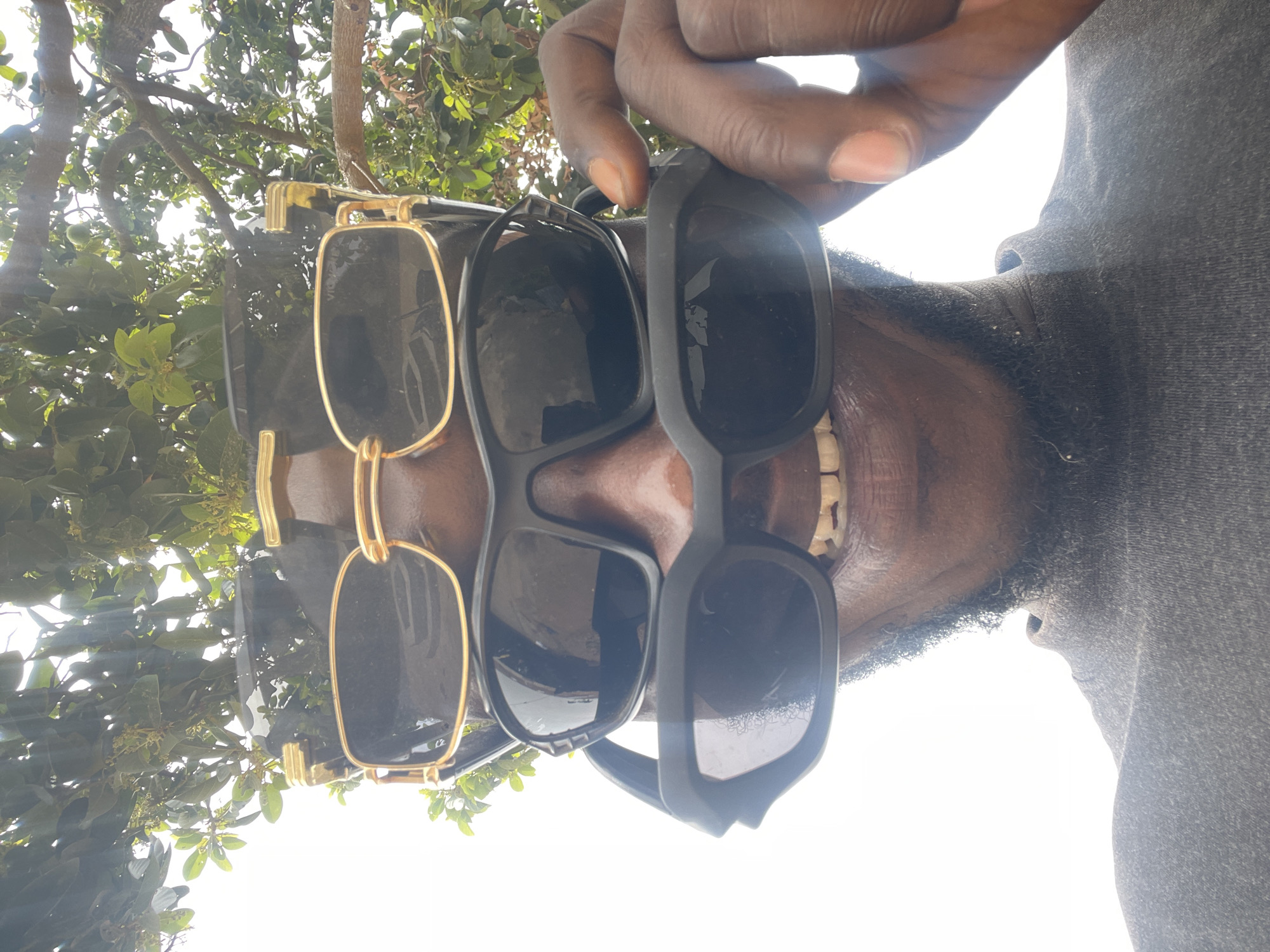 The image depicts a man with dark skin and a beard wearing three pairs of sunglasses, stacked on top of each other, while outside with trees overhead. No specific location in Jos, Nigeria, is discernible from the image.