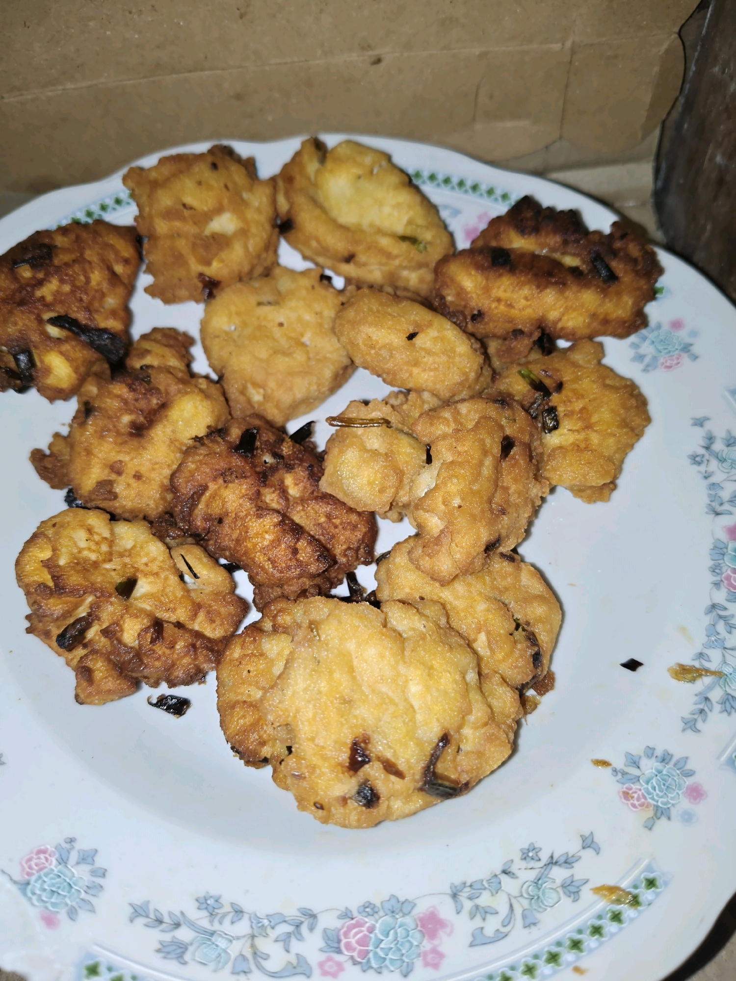 A plate of golden-brown fried fritters is piled high on a white plate with a floral pattern. The fritters appear to be made with chopped vegetables, possibly onions or scallions, mixed into a batter and deep-fried. They are irregular in shape and size, some with a slightly crispy, uneven texture. The plate is set against a neutral background that appears to be a textured wall or surface, suggesting an indoor setting. The lighting is dim, casting shadows and highlighting the texture of the food. The scene is likely from a home kitchen or a small eatery in Sumberwono, Indonesia, where such homemade fritters are a common snack. There are no people visible in the image.