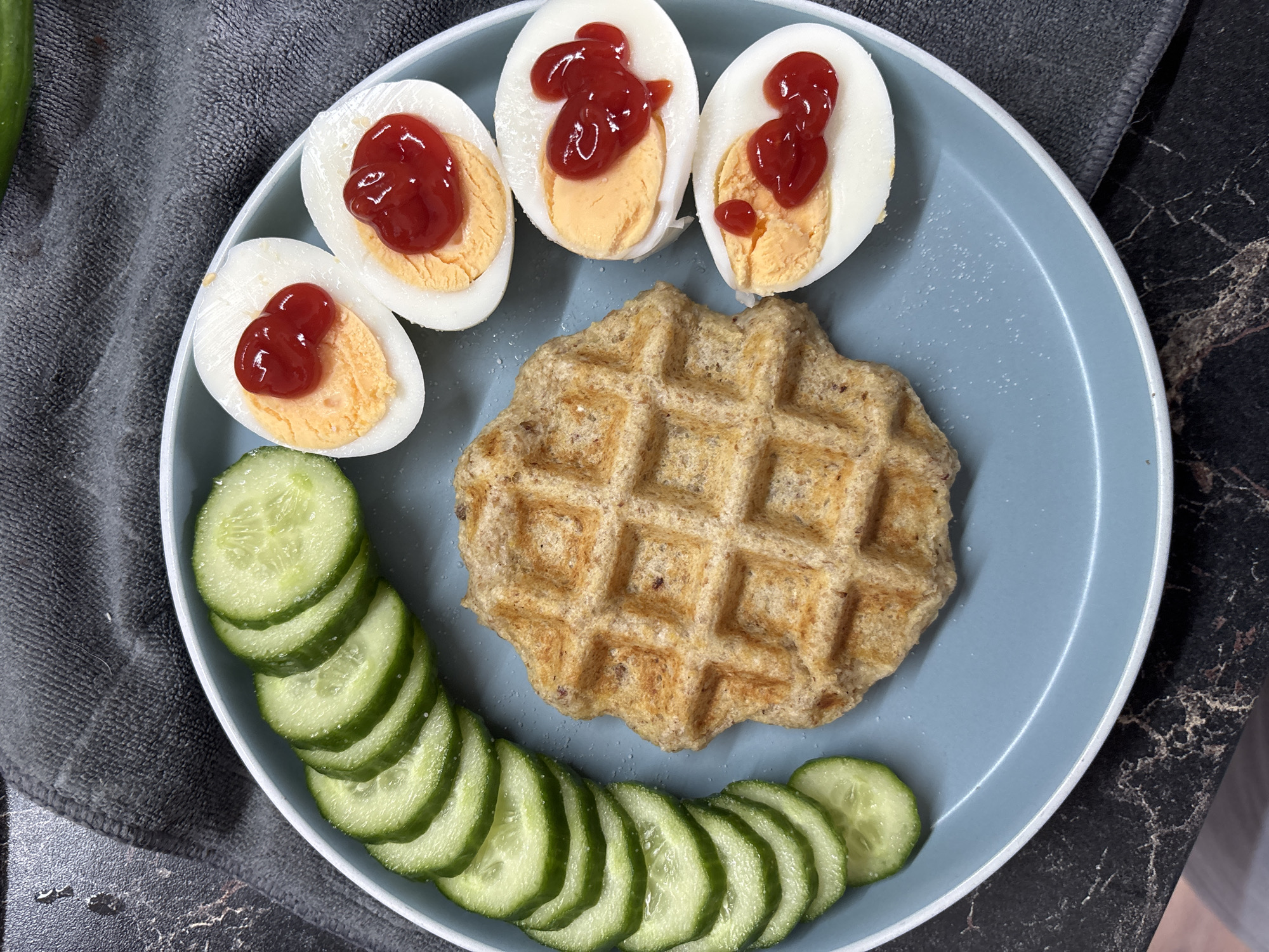A plate of food is centered on a dark, marble-patterned surface. The plate is light blue with a white rim. On the plate, there is a single, square waffle with a grid pattern, placed slightly off-center to the right. To the left of the waffle, there are several thin, circular slices of cucumber arranged in a curved line. To the right of the waffle, there are four halves of hard-boiled eggs, each topped with a dollop of ketchup. The eggs are arranged in a vertical line. A dark grey fabric napkin is visible behind the plate. The image is taken from a top-down perspective, with overhead lighting. There is no indication of the city of Sochi, Russia, or any people in the image.