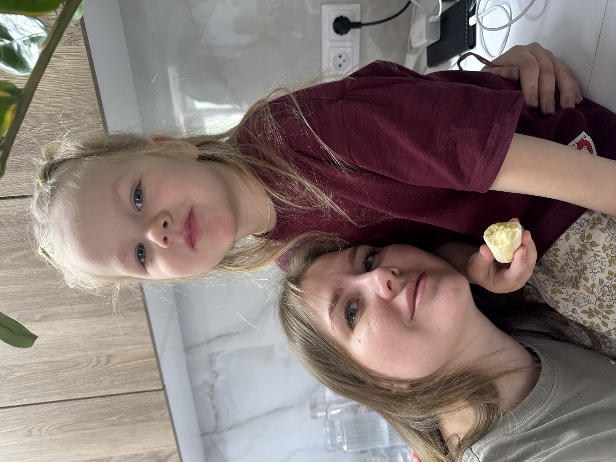 A photograph depicts a young girl and a woman, likely a mother and daughter, indoors. The girl has blonde hair styled with small hair bands, and she is wearing a burgundy shirt. The woman, who is holding a peeled banana, has light brown hair. A plant is visible in the foreground. Electrical outlets and cords are present on the wall behind them. The location appears to be inside a home, possibly a kitchen, given the cabinets. No specific details confirm the location is in Gornyi (Dvurechenskoe S/P), Russia.