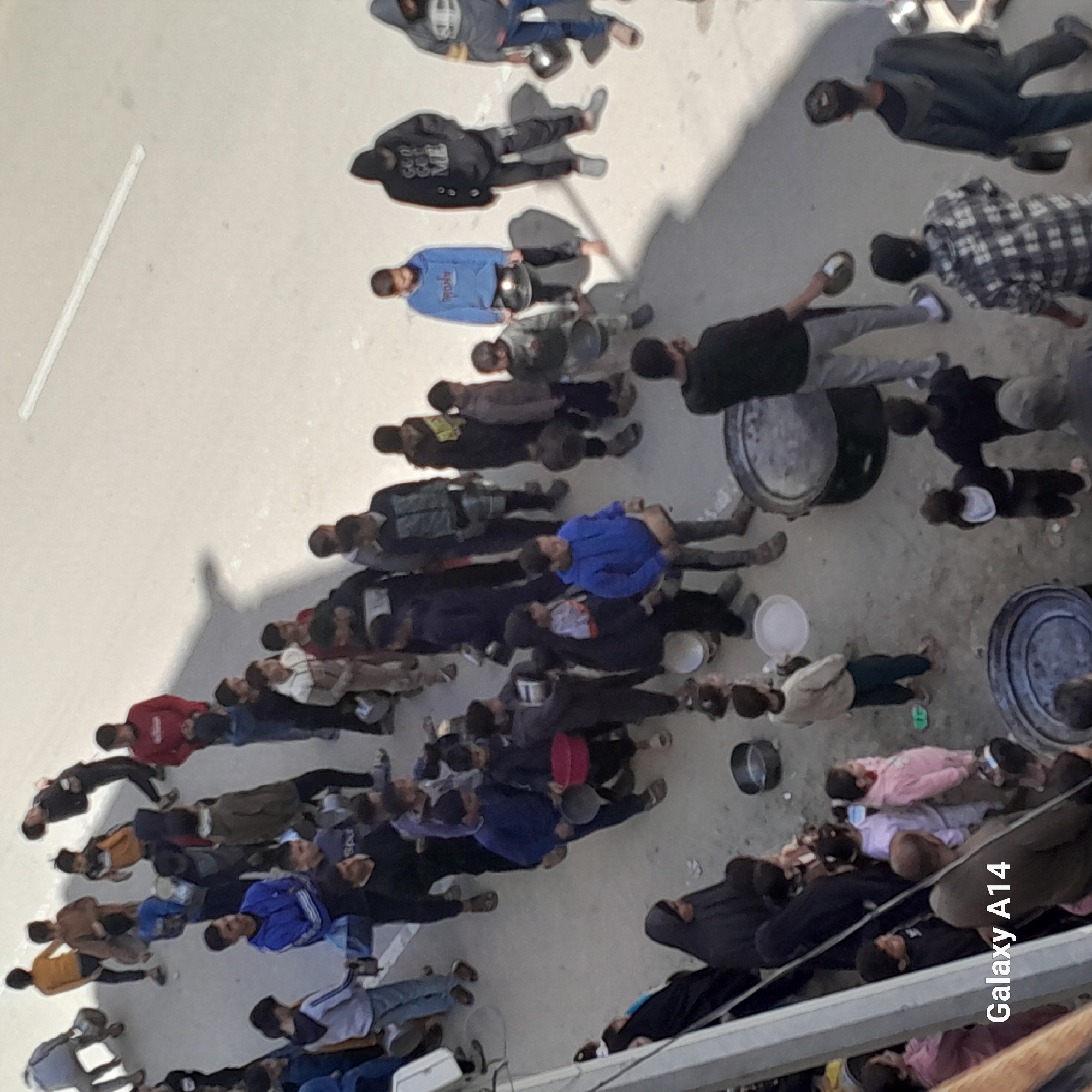A high-angle photograph depicts a large group of people gathered outside in what appears to be an urban environment. They are dressed in various casual clothing. Many of them are seated or lying on the ground, seemingly organized in rows or clusters. Some individuals are holding bowls or plates, suggesting they might be receiving or consuming food. There are metal containers of various sizes scattered around, possibly containing food or water. The ground is a light gray color, possibly concrete or pavement. A white line marking is visible in the upper left corner of the image. The location appears to be within the city of Unknown, Unknown.