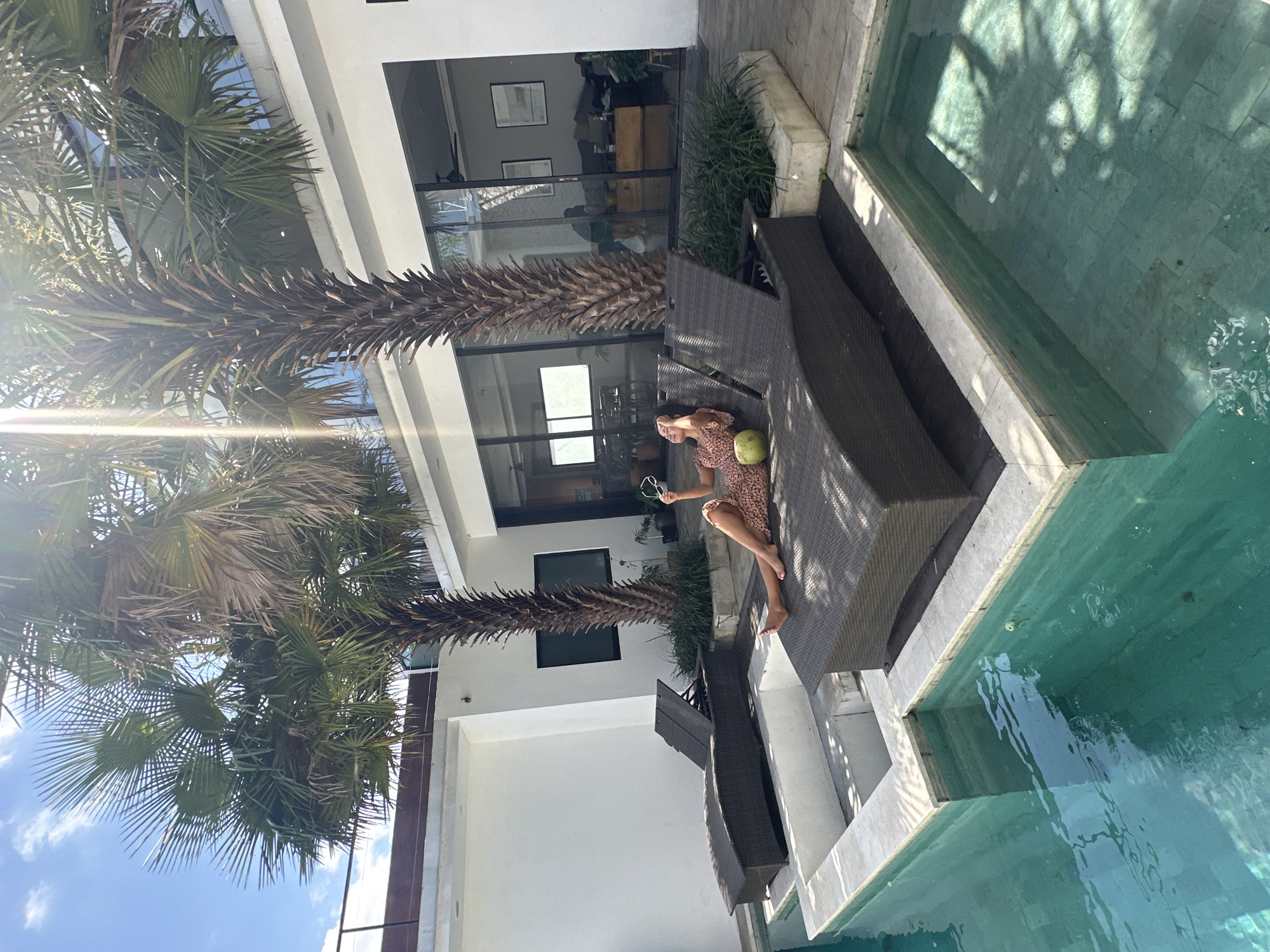 A woman in a patterned dress reclines on a dark brown lounge chair next to a turquoise swimming pool. She is holding a pair of sunglasses and a green coconut. Palm trees with large fronds are visible in the foreground, casting shadows on the scene. The background shows a modern building with large glass windows and white walls. The sky is blue with scattered clouds, suggesting a sunny day. The overall ambiance is one of relaxation and leisure, likely in a tropical resort or private villa setting in North Kuta, Indonesia. The lighting indicates it is daytime, possibly late morning or early afternoon.