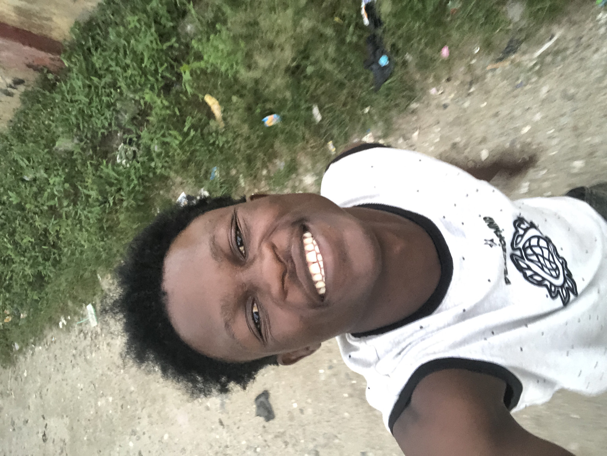 A young Black male, with short, dark, curly hair and dark brown eyes, smiles widely while looking directly at the camera. He is wearing a sleeveless white t-shirt with black trim around the neckline and armholes, and a graphic design on the front. He appears to be outside, with a dirt and gravel path to his left and a patch of green grass with some scattered debris on his right. The setting resembles an outdoor area within a city, potentially in Ado, Nigeria, given the presence of common urban elements like worn pathways and patches of unkempt vegetation. The lighting suggests it is daytime, possibly overcast or late afternoon, as there are no harsh shadows.