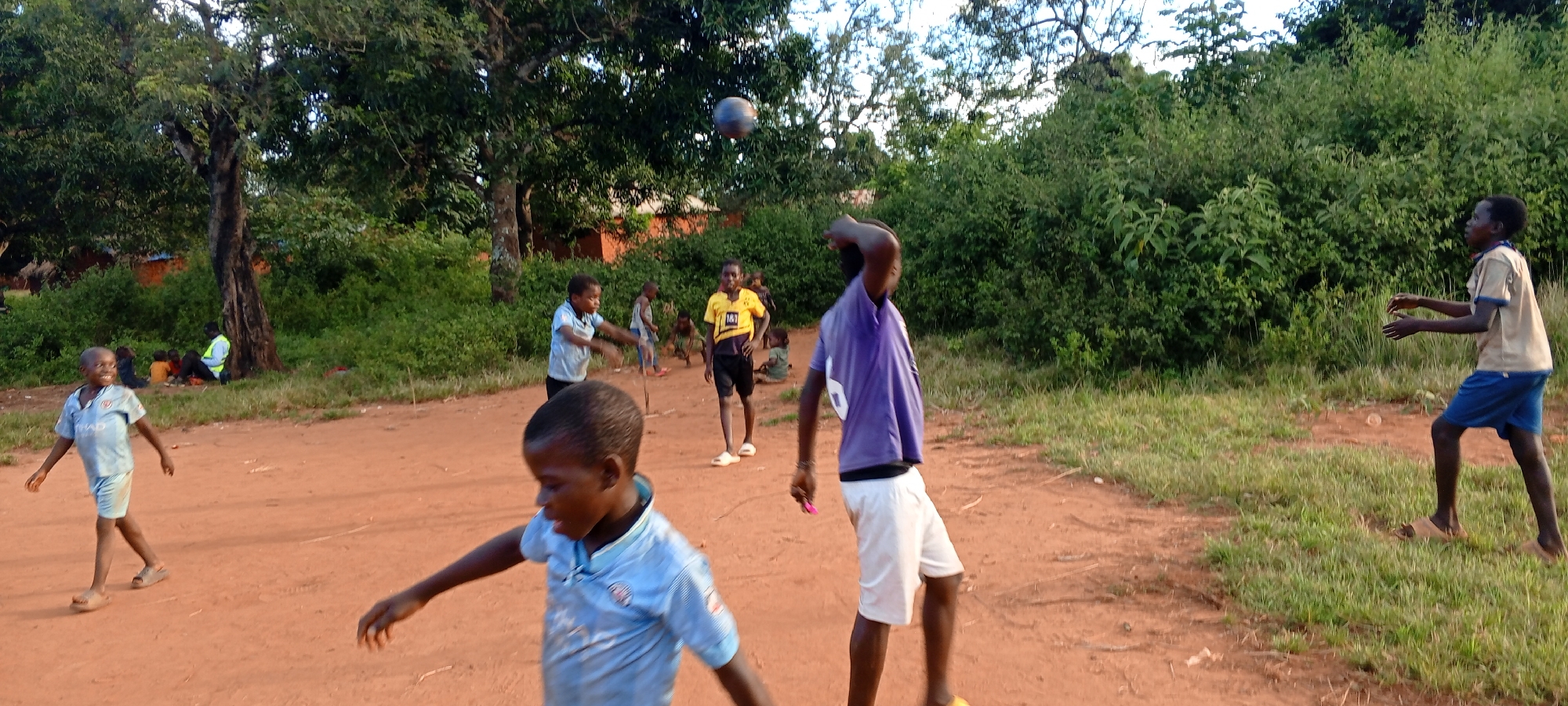 The image depicts a group of children playing in Ngalula, Democratic Republic of the Congo. The scene is set on a red dirt road, with trees and lush green bushes forming the backdrop. The sky is partly cloudy. There are seven children in the image, all wearing different clothing. One child is in the foreground wearing a light blue shirt. To his left a child is also wearing a light blue shirt and running. A child wearing a yellow Borussia Dortmund jersey and black shorts stands further away, and to his left, two more children are present. A child in a purple shirt and white shorts stands with a hand over his head, as if attempting to catch a ball. A black ball is in the air above him. To the right of the image, another child wears a light brown shirt and blue shorts. In the background, other children are sitting near a tree. A man in a bright yellow vest is nearby.