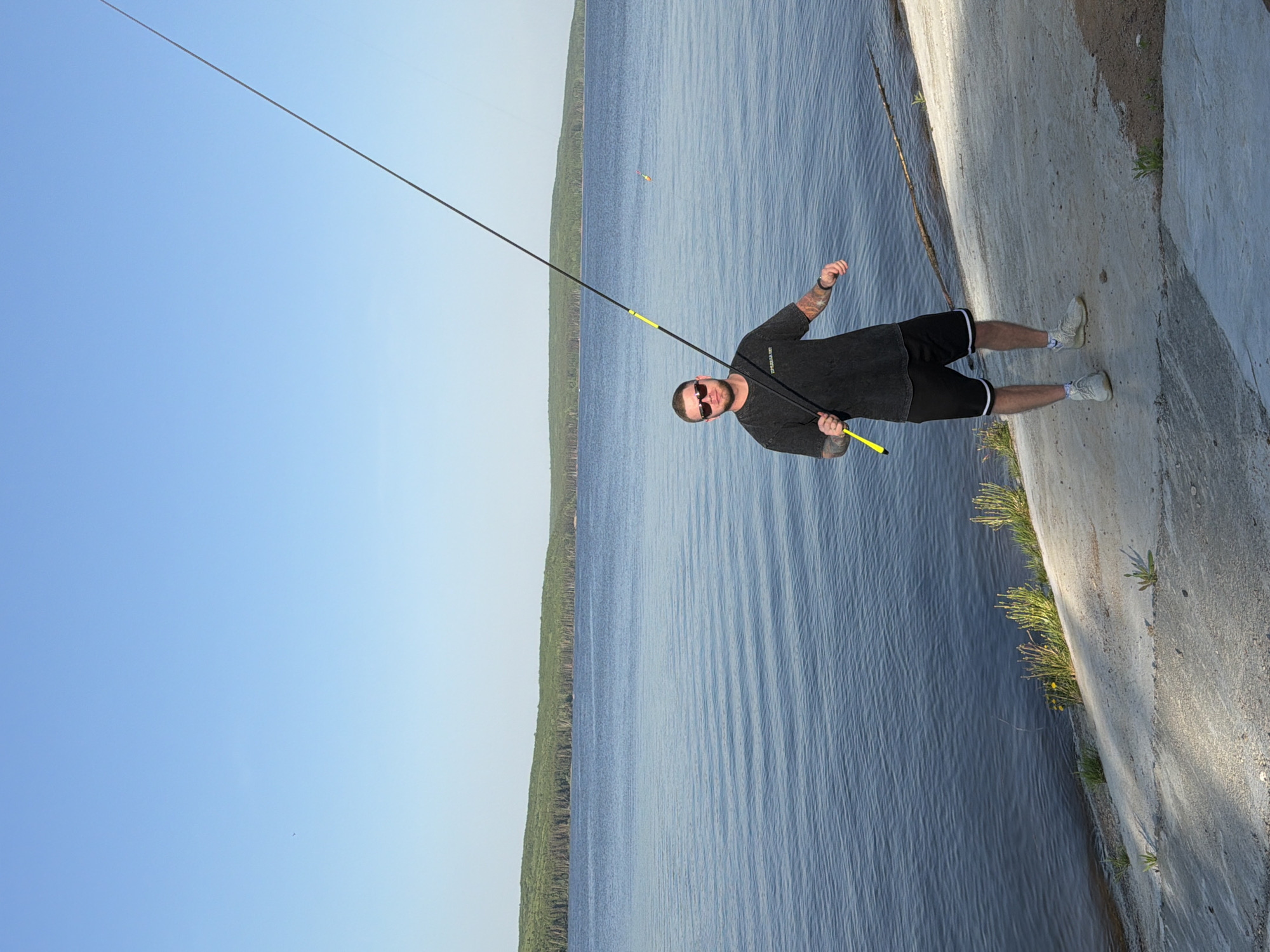 A man wearing sunglasses, a gray shirt, and black shorts with white trim stands on a concrete embankment holding a fishing rod. The location is Chaikovsky, Russia. He is fishing in a large body of water with a treeline visible on the opposite shore. The sky is clear and blue. There is a float visible in the water. Grass and vegetation grow on the embankment.