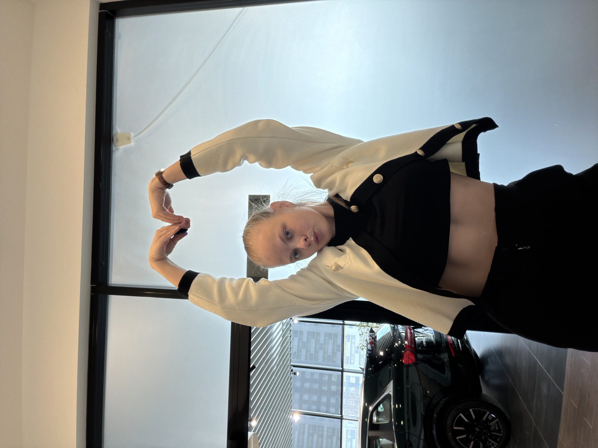 A young woman poses for a photograph indoors, forming a heart shape with her hands and arms above her head. She looks directly at the camera with a neutral expression. She wears a black crop top, black trousers, and a cream-colored cardigan with black trim and prominent gold buttons, along with a brown-strapped watch on her left wrist. The setting features large, black-framed glass windows that overlook an urban landscape with reflections of buildings and a light, possibly overcast, daytime sky. In the lower right background, the rear section of a dark-colored SUV is visible, including a wheel and taillight; faint text on the car suggests it might be a 'SKODA' model. A plain white wall borders the left side of the frame. This scene is located in Izhevsk, Russia.