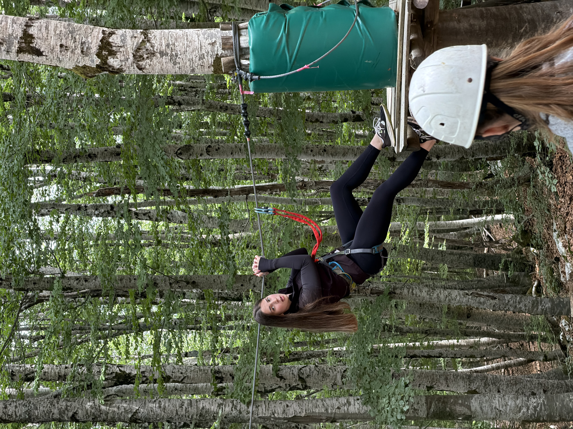 The provided image, rotated 90 degrees counter-clockwise, depicts a person engaged in an outdoor adventure activity within a dense forest at Dolovi, Montenegro. A young woman, the central subject, is suspended downwards from a safety harness and red tether, attached to a zip-line cable or high ropes course element. She wears black athletic attire, including a long-sleeved top, leggings, and black sneakers with white laces, and looks directly at the camera with a calm expression. Her long brown hair falls freely.
Above her, partially visible to the right, is another individual wearing a white hard hat with "VIP" printed on its strap. The zip-line setup includes a grey cable connected to a large green padded cylinder mounted on a wooden structure, likely a platform. The surrounding environment is characterized by numerous tall trees with light-colored, patterned bark and abundant green leaves, indicative of a lush natural setting. The scene is brightly lit, suggesting it is daytime during pleasant weather conditions.