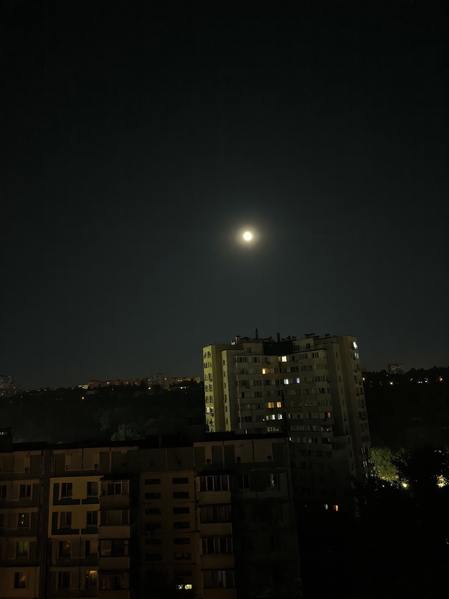 The image depicts a night scene in the city of Chișinău, Moldova, with a prominent full moon illuminating the sky. In the foreground, two apartment buildings are visible. The closer building on the left is partially in shadow, revealing several lit windows. The building on the right, further back and taller, is largely illuminated by the moon and has many windows, some of which are lit with warm yellow and orange light. Behind these buildings, a dimly lit cityscape stretches into the distance, with outlines of more buildings and trees silhouetted against the dark sky. The overall atmosphere is quiet and urban, with the moonlight casting a soft glow over the scene.