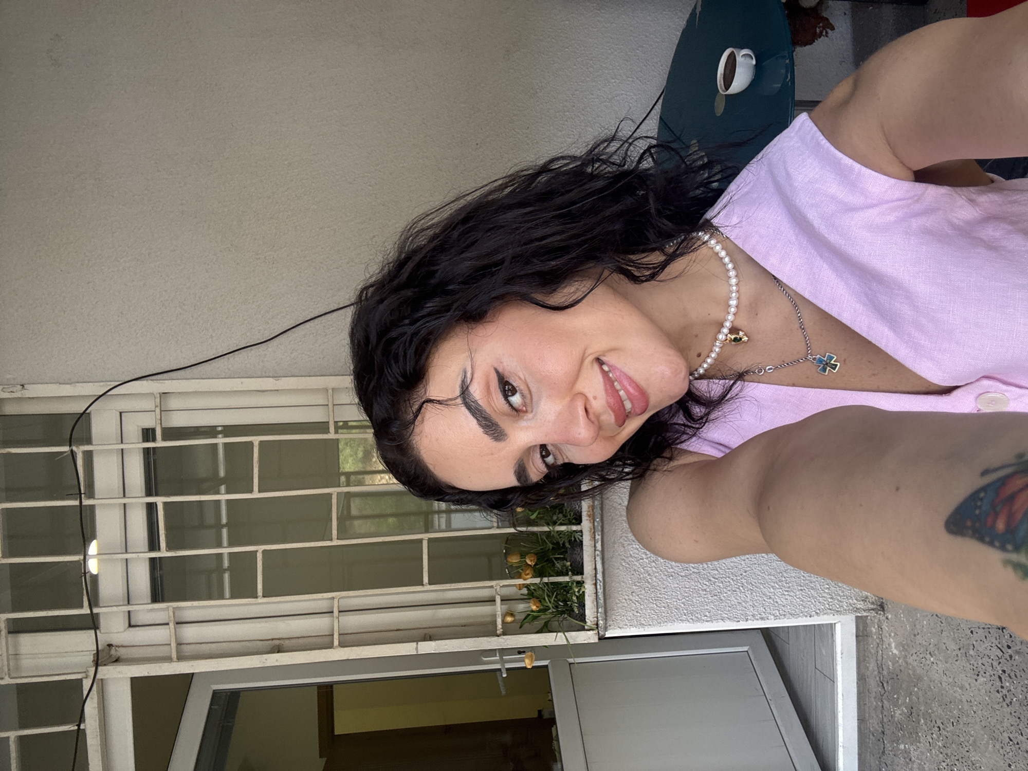 A young woman with dark, curly hair and fair skin is taking a selfie outdoors. She is smiling and looking at the camera. She is wearing a light pink, sleeveless, V-neck top and several necklaces, including a pearl necklace with a small green charm and a silver chain with a blue and green cross pendant. Her left arm is extended towards the camera, and a tattoo of a butterfly is visible on her upper arm.
The background shows a building facade with a white door and a window with white bars. In front of the window is a planter box with several yellow tulips. To the right of the woman, a dark green table with a small white cup of coffee is visible. The overall setting appears to be a courtyard or balcony area. The image has a bright, natural light, suggesting it was taken during the daytime. The presence of tulips suggests a spring or early summer setting. The architecture of the building and the style of the window bars are consistent with residential buildings found in Tbilisi, Georgia, particularly in older neighborhoods.