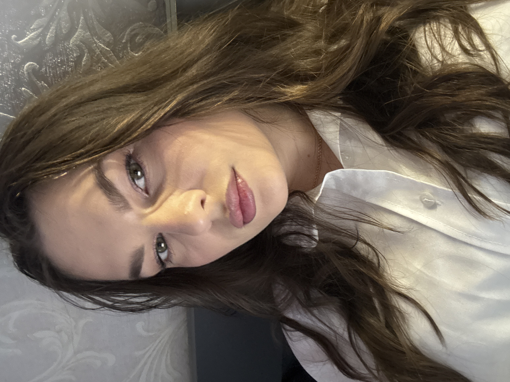 A young woman with brown hair and green eyes is posing for a photograph. She is wearing a white button-down shirt. The background appears to be a textured wall. The photo might have been taken indoors, but there is nothing to link it to the city of Chaikovsky.