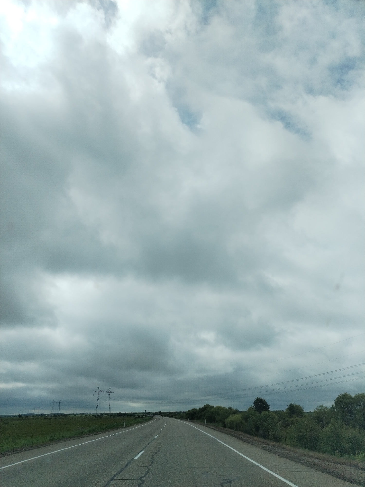 The image shows a desolate, overcast landscape with a paved road leading into the distance. Power lines stretch across the horizon, supported by tall, skeletal towers. The road is lined with sparse vegetation and bushes. The sky is filled with dramatic, heavy clouds, suggesting an impending storm or a post-storm gloom. There are no people or vehicles visible in the scene. The overall impression is one of isolation and a vast, open, but somber environment. The location is identified as Unknown, Unknown.