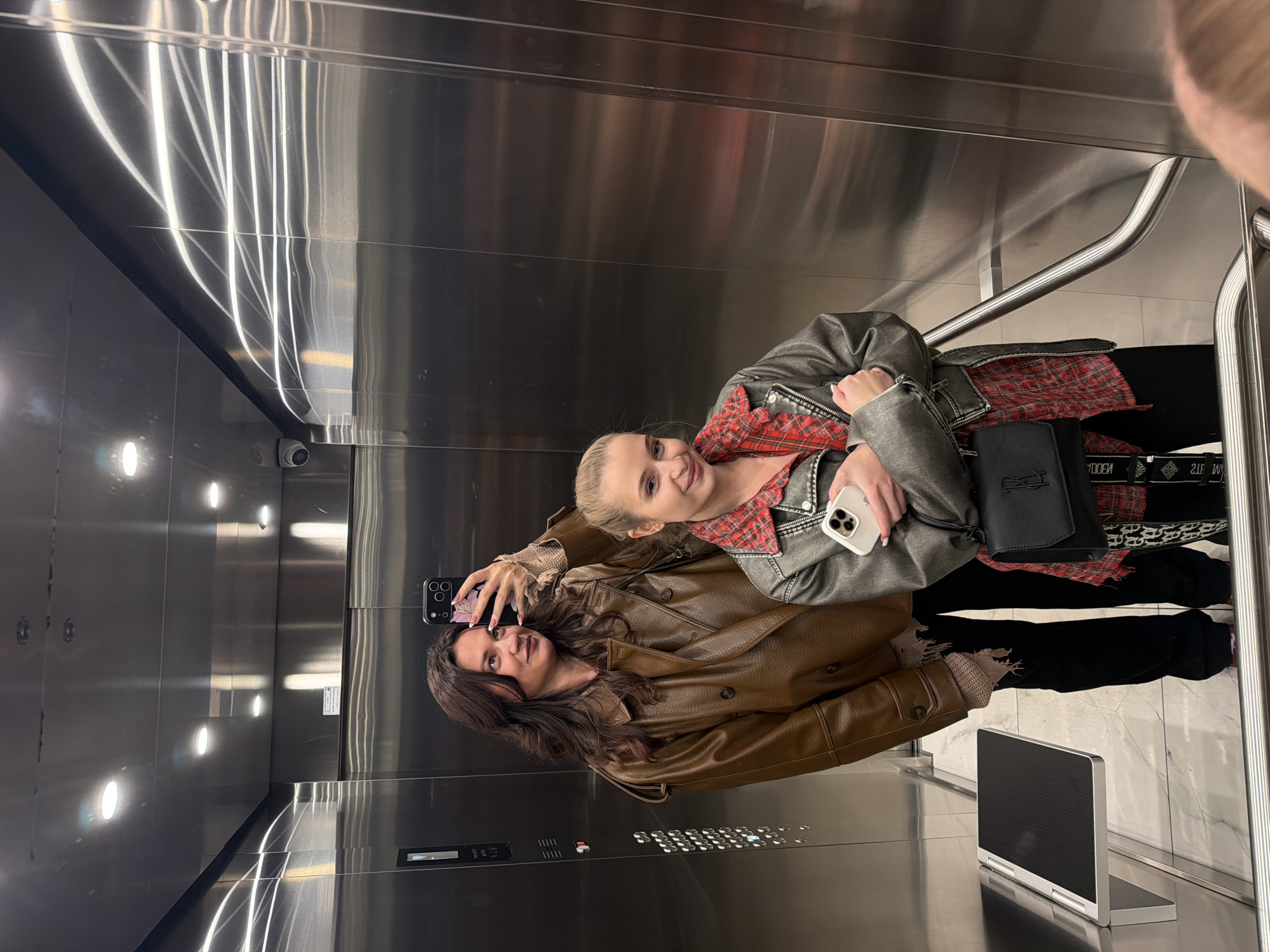 Two women take a selfie in an elevator with stainless steel walls. The elevator is equipped with a handrail and a digital display panel. One woman has long brown hair and wears a brown leather jacket. The other woman has blonde hair and wears a grey leather jacket over a red plaid shirt. A black purse rests against the handrail. The location is Belgrade, Serbia.