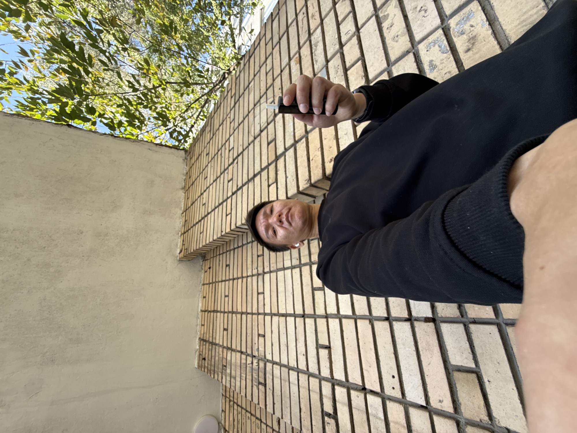 In Moscow, Russia, a low-angle shot shows a man holding a black electronic cigarette in his right hand, positioned in front of a brick wall with a slight upward tilt. The man is wearing a black sweatshirt and white earbuds. Above the brick wall, a portion of a light-colored concrete structure and green foliage under a bright blue sky are visible. The image is captured outdoors during the day.
