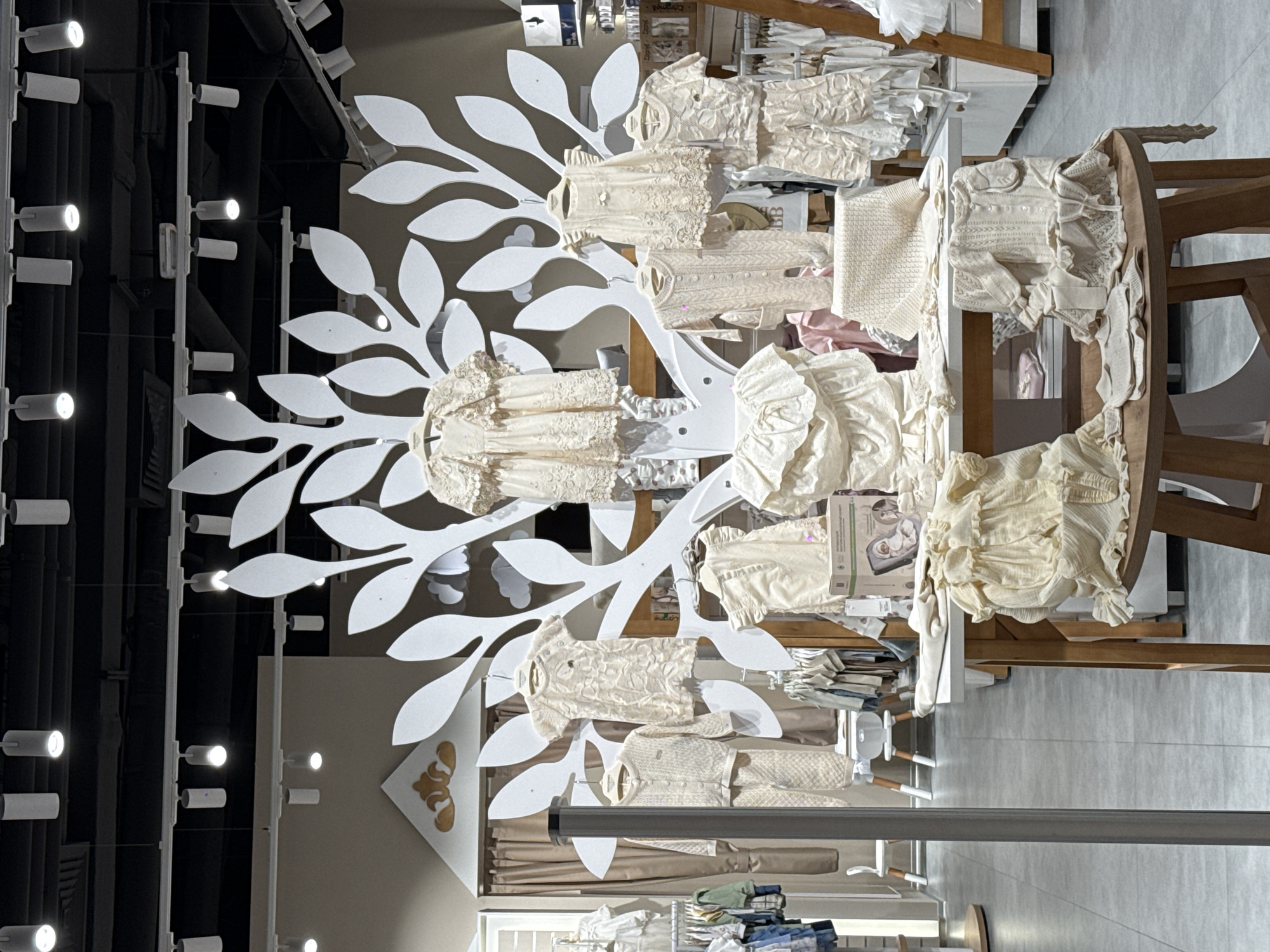 A retail display is photographed at an upward angle. A white, decorative tree structure with leaf cutouts displays beige children's clothing. The clothes are neatly arranged on the tree's "branches." The tree is positioned in front of a wall with a decorative emblem. A clothing rack is in the background, holding various garments. The ceiling is visible, featuring track lighting. A metal pole, likely part of the store's structure, runs horizontally across the lower part of the frame. The setting appears to be a children's clothing store in Moscow, Russia.