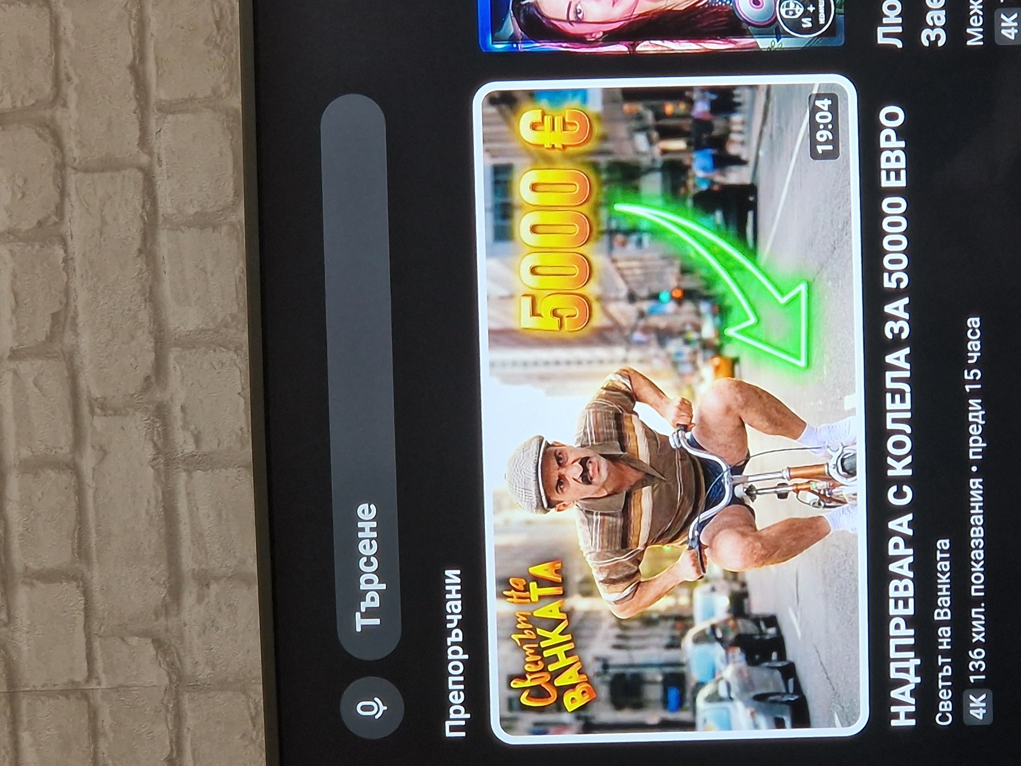 A screen displays a digital interface, likely a video streaming platform, against a background of a light brown, textured brick wall. The screen shows a dark grey top section with a search bar featuring a white microphone icon and the Cyrillic text "Търсене" (Search). Below this, the Cyrillic text "Препоръчани" (Recommended) is visible.
The central focus is a video thumbnail bordered in white, depicting a light-skinned man with a dark mustache and a stern expression. He wears a brown and white striped polo shirt and a grey plaid flat cap, and is actively riding a small bicycle. His legs, clad in white socks and shoes, are bent high as he pedals. Overlay text in the top left of the thumbnail reads "Светът на ВАНКАТА" (The World of VANKATA) in orange/yellow letters, while "5000 €" in large yellow text appears in the top right, accompanied by a bright green upward-curving arrow graphic. The background of the thumbnail shows an urban street scene with vehicles and buildings, representing a city environment of Unknown, Unknown. A timestamp "19:04" is visible in the bottom right of the thumbnail.
Below the thumbnail, the video's title is displayed in white capital letters: "НАДПРЕВАРА С КОЛЕЛА ЗА 50000 ЕВРО" (RACE WITH BICYCLES FOR 50000 EURO). The channel name, "Светът на Ванката" (The World of Vankata), is beneath the title. Further details state "4K 136 хил. показвания • преди 15 часа" (4K 136 thousand views • 15 hours ago), indicating resolution, view count, and upload time. A portion of an adjacent video thumbnail showing a woman's face is visible to the right.