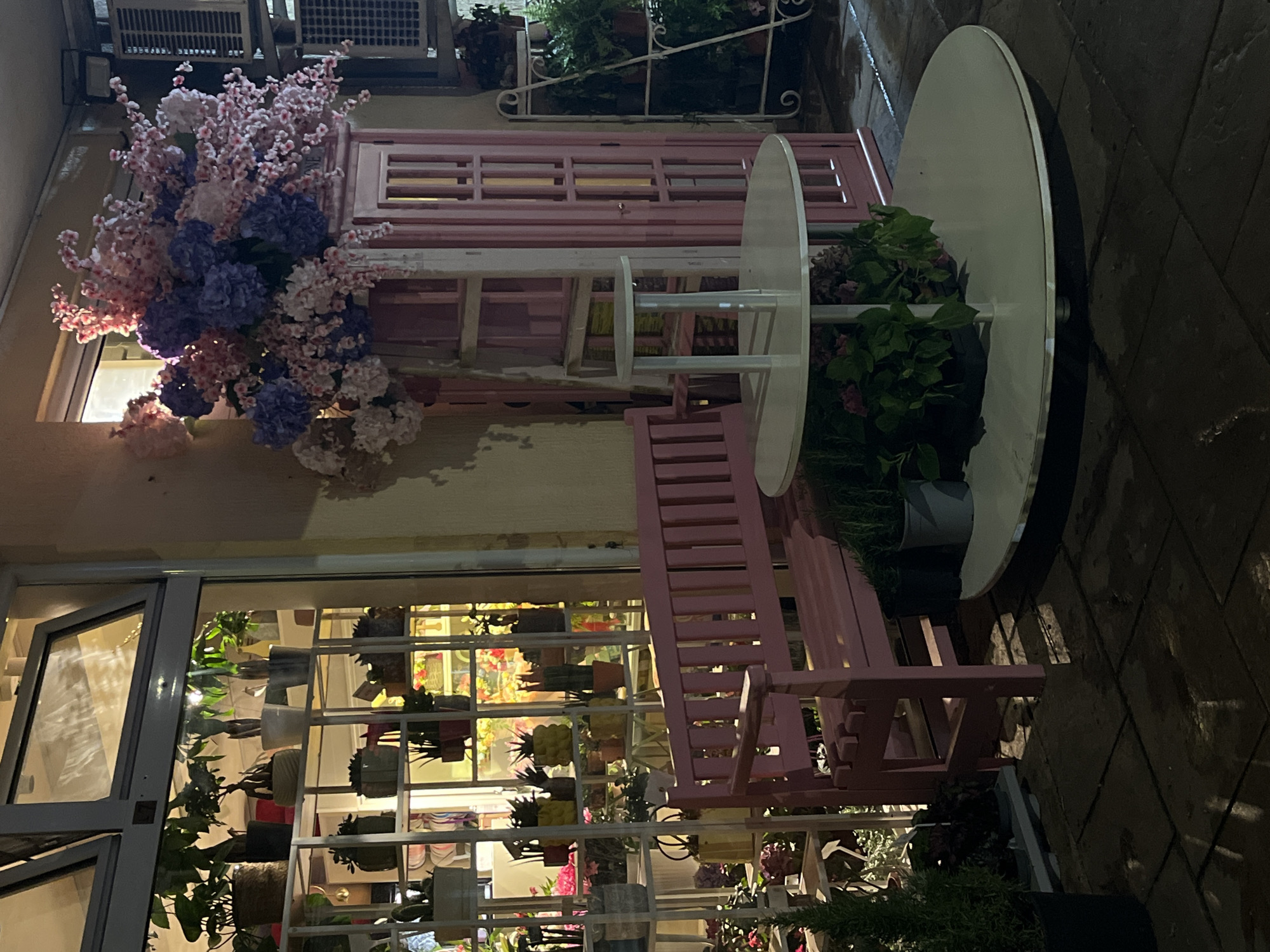 The image depicts an outdoor seating area in front of a shop, possibly a flower shop, in Budva, Montenegro. The scene is dimly lit, suggesting it is either nighttime or very early morning. The shop window on the left displays numerous potted plants on white shelving units. To the right of the shop window is a pink wooden bench. In front of the bench are two round, tiered tables with a lighter colored top tier and a darker base. Behind the tables and bench, there is a pink cabinet with glass-paneled doors, adorned with a large arrangement of artificial pink and purple flowers at the top. The ground is paved with dark rectangular tiles, some of which appear wet. To the far right, a white metal shelving unit holds potted plants. The overall impression is one of a charming, somewhat whimsical, outdoor display. There are no people visible in the scene.