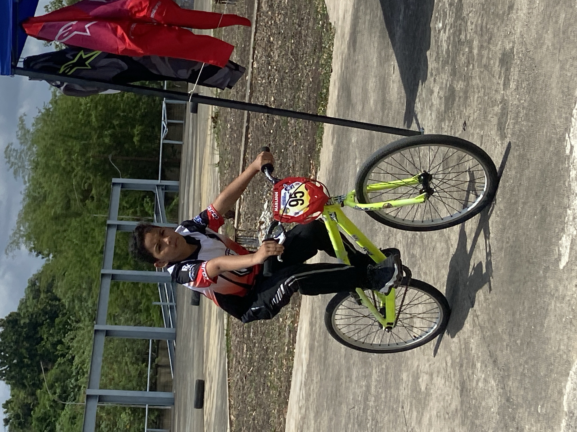 A young person rides a yellow BMX bike on a concrete surface. The person wears a red, white, and black jersey, black pants with a white stripe, and black shoes. The bike has a red number plate with the number 95. The name "MARIANN" is visible on the number plate. In the background, there are trees, a metal structure, and clothes hanging on a stand. The location is Tinaquillo, Venezuela.