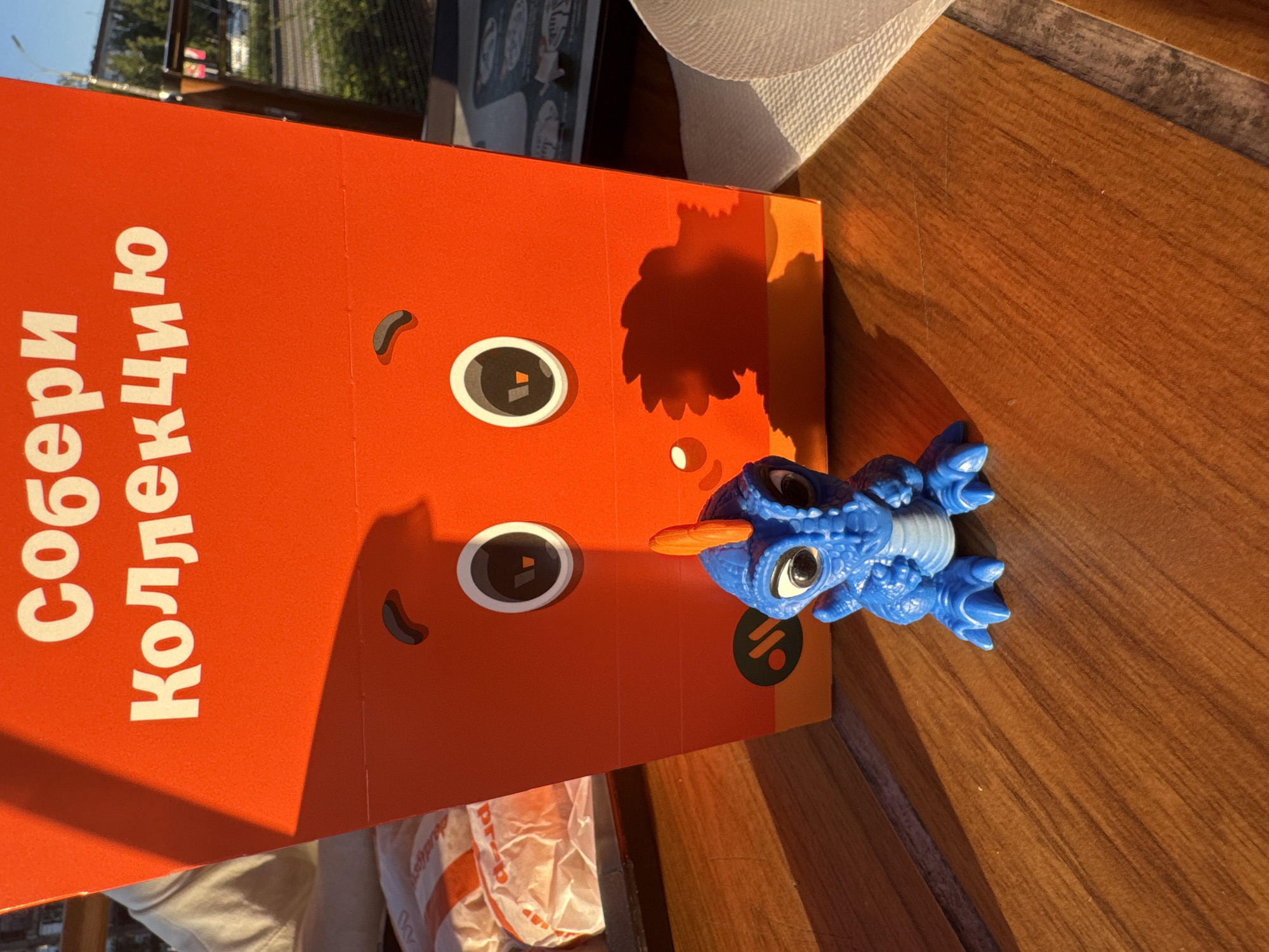 This image captures an outdoor dining scene at a fast-food establishment in Izhevsk, Russia. A vibrant orange fast-food meal box, adorned with white Cyrillic text "Собери Коллекцию" (Collect the Collection) and cartoonish facial features (eyes and a mouth/nose), dominates the center. A small, blue, reptilian-like collectible toy with an orange head crest and black eyes stands directly in front of the box on a polished brown wooden table. Intense direct sunlight illuminates the scene from the left, casting prominent shadows from both the box and the toy onto the table surface. A person's light-colored arm and shoulder are partially visible behind the left side of the box. To the left, a crumpled white plastic bag containing additional food items is visible. A white textured napkin rests on the table to the right. The background shows an urban environment with a visible street, distant trees, and indistinct buildings under a bright sky. The branding on the box, including a stylized "V" in the eyes and a green circular logo, is consistent with Vkusno i Tochka.