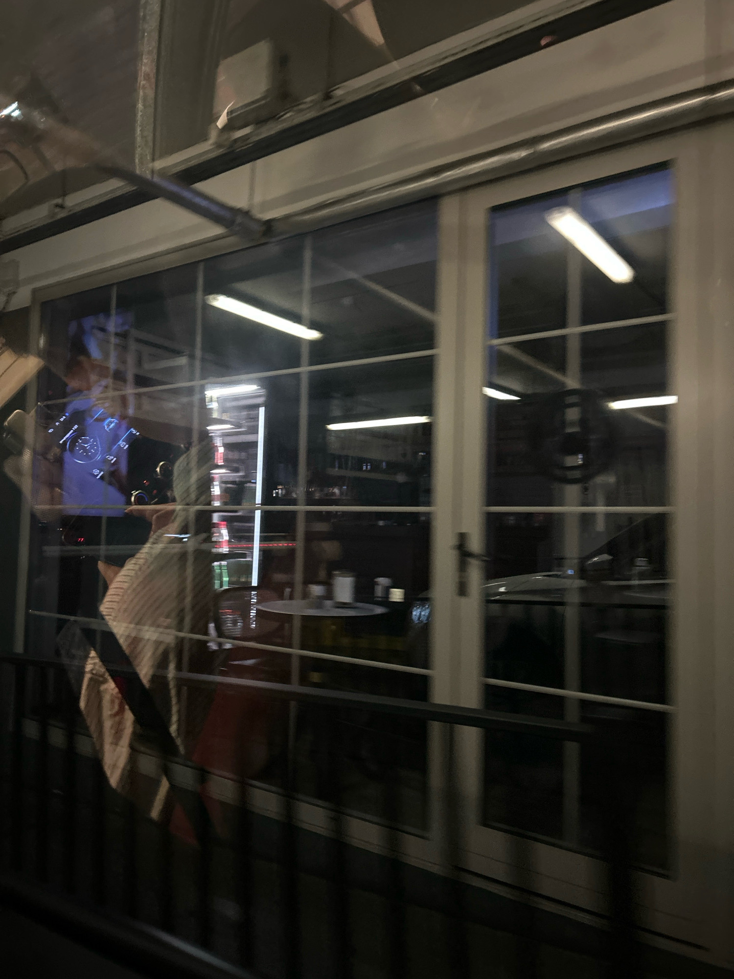 The image is a blurry, night-time, interior shot through a window, with reflections distorting the view. The scene appears to be inside a building, possibly a restaurant or cafe, with glass doors and windows. Fluorescent lights are visible, some appearing as streaks due to motion blur. In the foreground, a dark metal railing runs horizontally across the bottom of the frame.
Reflected in the glass are elements of an outdoor urban environment, including streetlights and what appear to be building facades. The interior shows tables and chairs, with some bottles visible on shelves in the background. A person wearing a striped shirt is partially visible, with their arm and shoulder illuminated.
The image's low light and motion blur make it difficult to discern specific details about the location. However, given the context of Baku, Azerbaijan, the architecture and lighting might be consistent with various establishments in the city, though no definitive landmarks are identifiable. The scene conveys a sense of movement and a transient glimpse into a nighttime establishment.