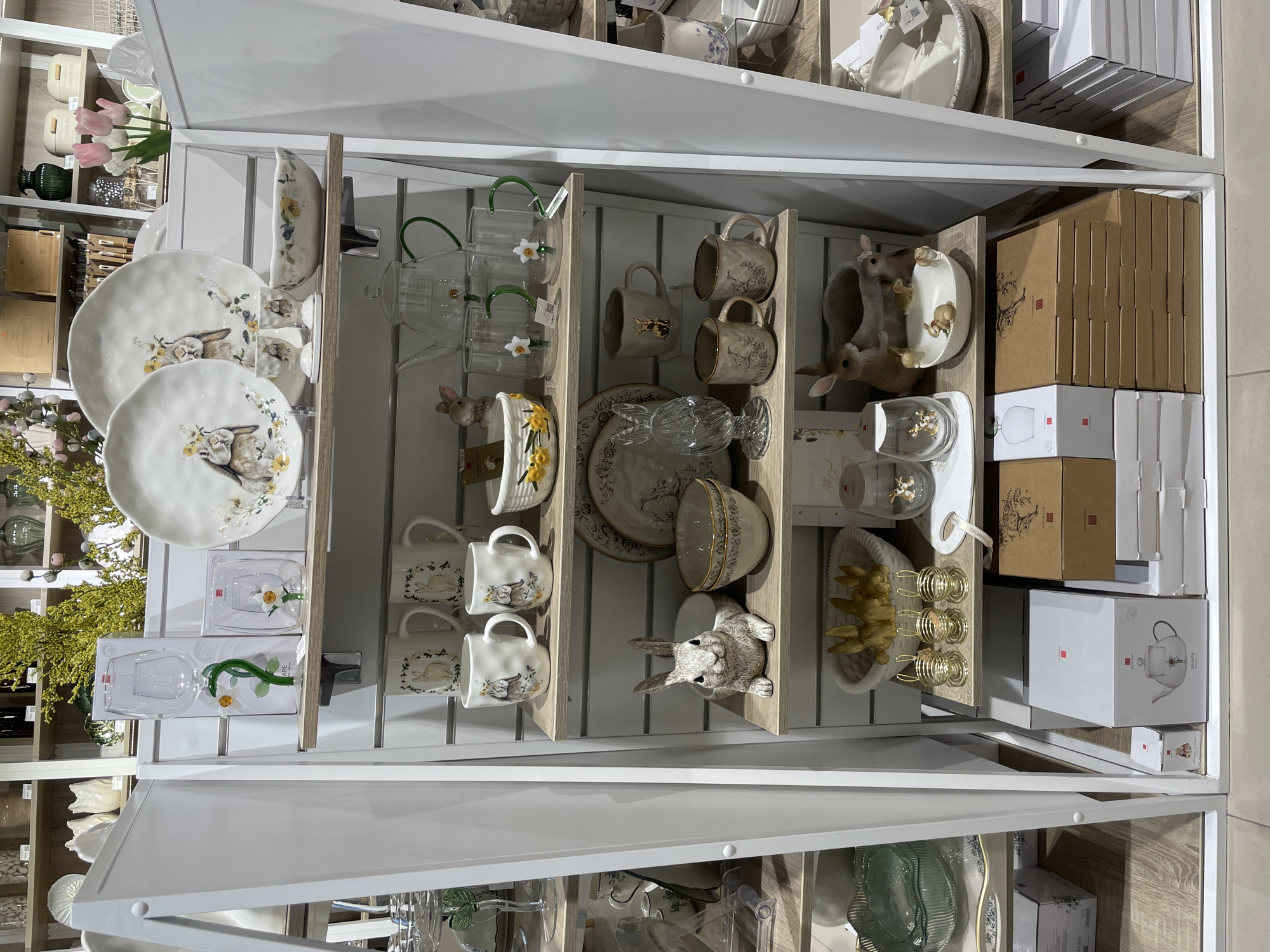 This media file is a photograph of a retail display shelf showcasing Easter-themed tableware. The scene is an indoor retail environment, likely a home goods store. The shelves are made of light-colored wood and white metal.
Prominently featured are ceramic plates and mugs adorned with illustrations of rabbits and floral motifs, typical of Easter decor. There are also glass pitchers with green handles and flower accents, and a decorative ceramic bunny figurine. Other items include bowls, boxes of merchandise, and various smaller decorative objects.
The overall impression is one of a neatly organized retail display, designed to appeal to customers looking for seasonal home decor and gifts. There is no indication of the time of day or weather. The location context is provided as Perm, Russia.
Visible text includes branding and product information on the packaging and labels, such as "Kitchencraft" on some of the boxes.