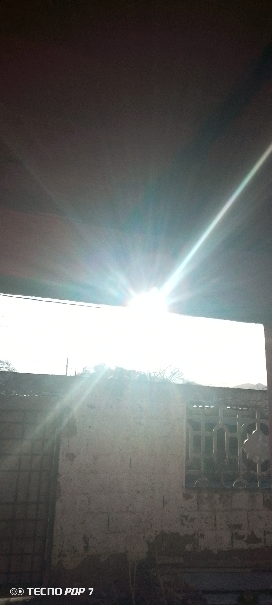 The image shows an exterior view in Altagracia, Venezuela, with a partially obscured view of the sky, dominated by bright sunlight. Visible elements include a section of a weathered, possibly brick, wall. A barred window is present, as is a metal gate. The ground in the foreground is unclear. The upper portion of the image is shadowed by an overhanging structure. A "TECNO POP 7" watermark is visible at the bottom left.