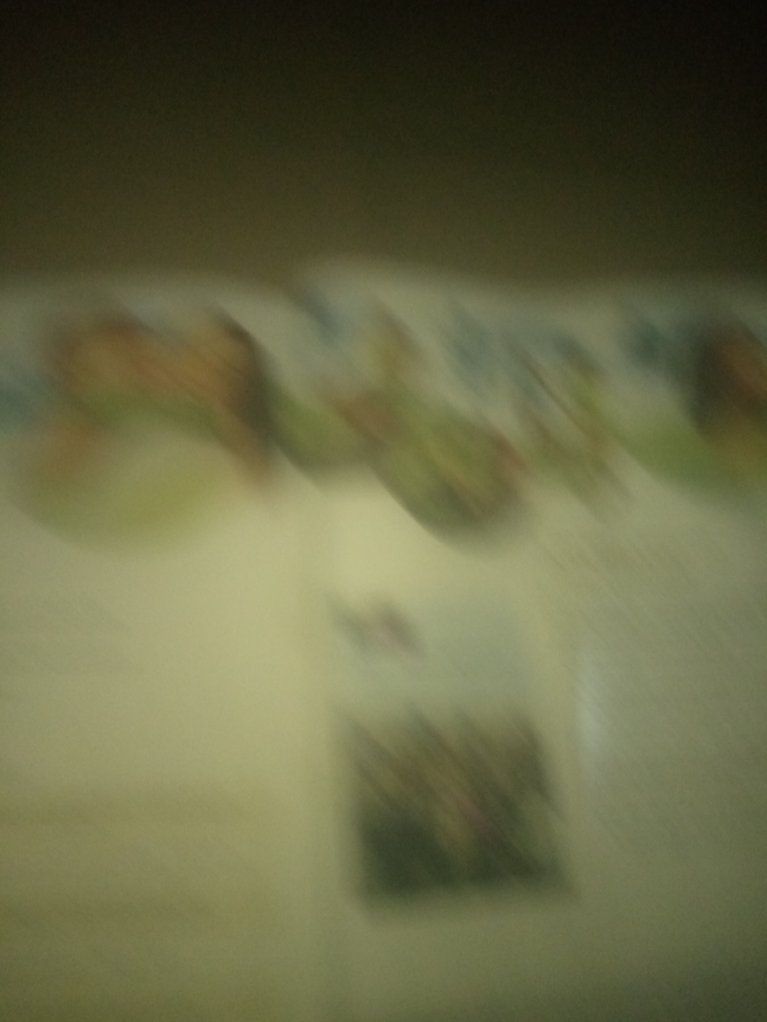 The image is an out-of-focus, low-resolution photograph of what appears to be a surface with some blurry objects or text on it. The top portion of the image is dark, transitioning to a lighter, yellowish-green hue towards the bottom. There are diagonal streaks of muted colors, predominantly greens, browns, and some lighter shades, suggesting movement or a textured surface. A small, dark, rectangular area with faint patterns is visible in the lower right quadrant. The image quality makes it impossible to discern specific details of any objects or discern any identifiable features within Jauro Yinu, Nigeria.