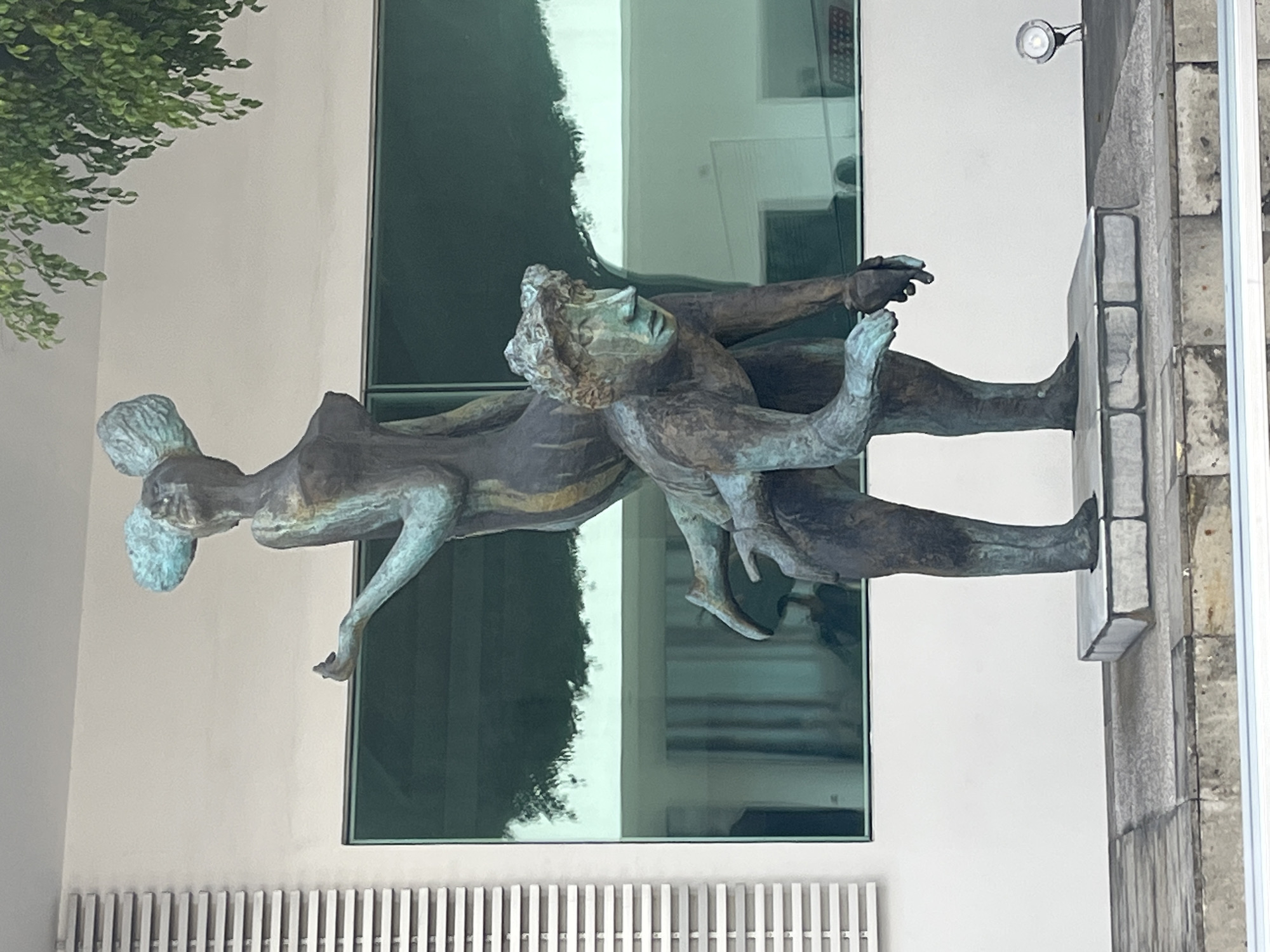 A bronze sculpture depicting two human figures stands outdoors against a modern building. The sculpture, rotated clockwise in the image, shows a larger, standing figure supporting or carrying a smaller figure. The larger figure has a robust build, short textured hair, and appears to be holding the second figure with both arms. The smaller figure is elevated and angled, with bent legs and head looking upwards, also featuring textured hair, possibly in a bun. The bronze material shows a prominent greenish patina, especially on the hair and body parts.
The sculpture rests on a dark, low base that connects to a textured stone or brick wall on the right. In the background, a light-colored building features large, dark, reflective glass windows, which show reflections of interior spaces and possibly parts of the surrounding environment. Green foliage from a tree or bush is visible in the upper left corner. A small, silver-colored security camera or light fixture is mounted on the building wall in the upper right. A metal grate or railing runs along the bottom of the image. The scene is illuminated by daylight, and no text is visible.