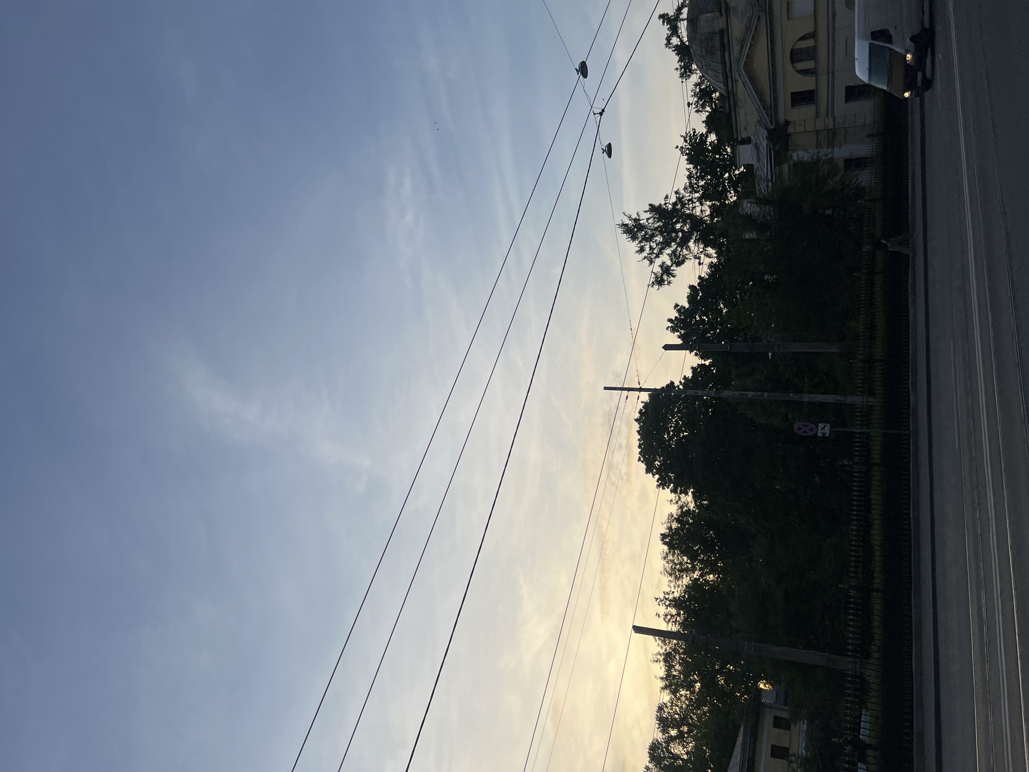 The image shows a street scene with a road in the foreground, and buildings and trees in the background, silhouetted against a twilight sky. Several overhead wires crisscross the frame, and some lampposts are visible. A white van is driving on the road to the right of the frame. The sky is a pale blue, with wispy clouds and a faint glow of sunset colors near the horizon. The overall impression is one of a quiet evening in an urban environment.
