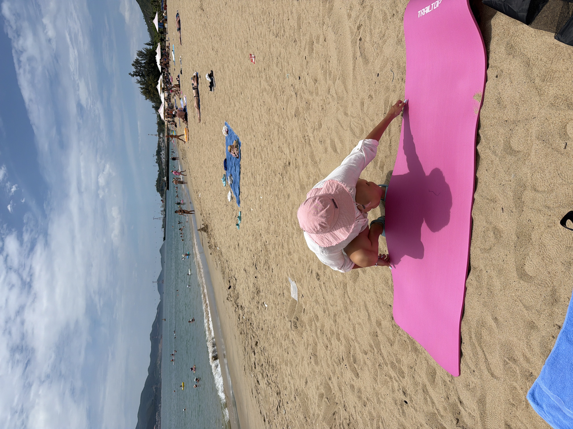 A young woman wearing a pink sun hat and a white shirt crouches on a sandy beach in Nha Trang, Vietnam, near the water's edge. She is in the process of unrolling a bright pink yoga mat. The beach is populated with other beachgoers sunbathing on towels and swimming in the ocean. Beach umbrellas are set up in the distance, and a lush, green mountain range forms the backdrop. The sky is partly cloudy, with a gentle breeze visible in the waves lapping at the shore.