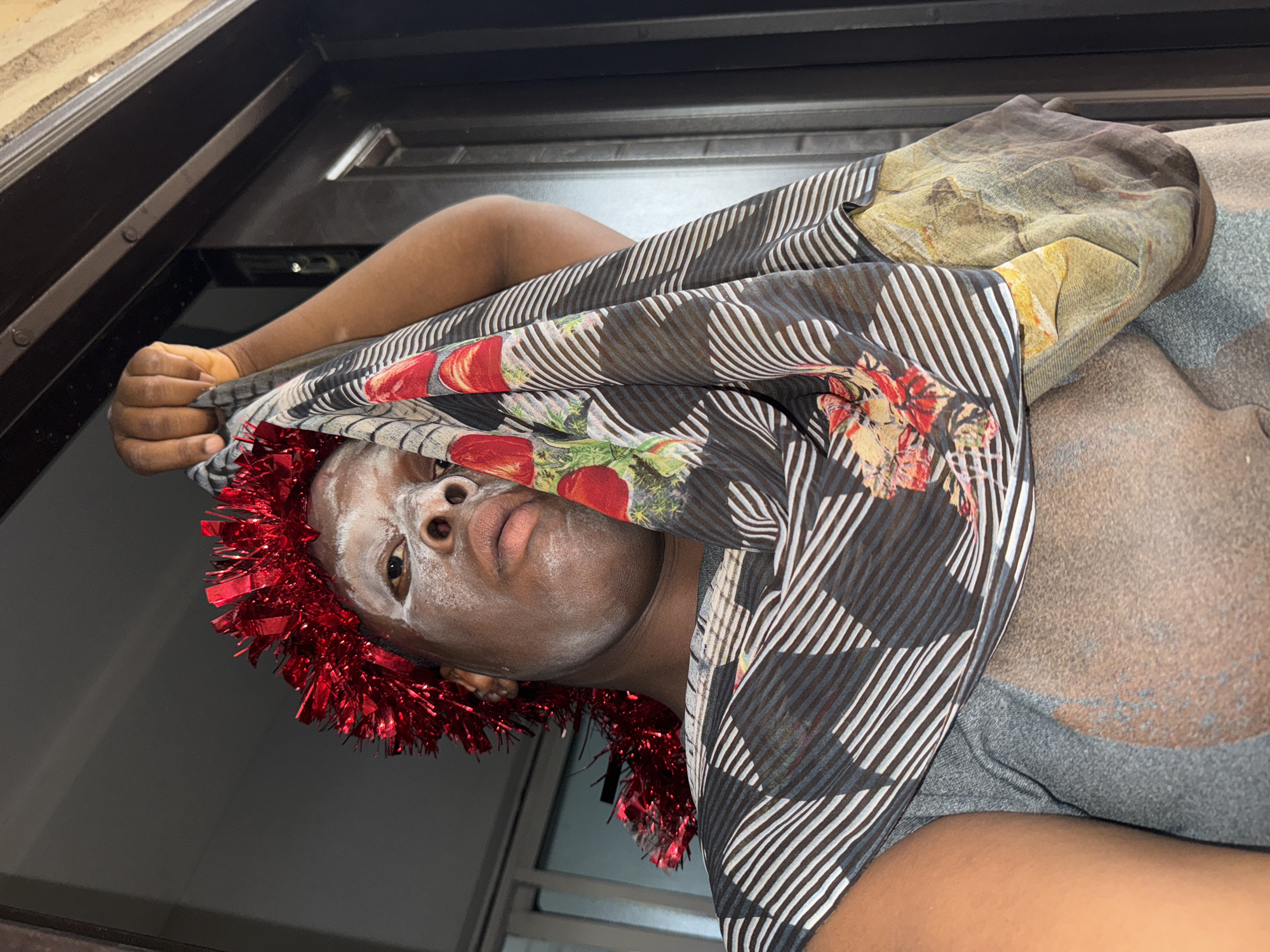 A person with a dark complexion is positioned in front of a window frame. The person's face is covered in a white substance, and a red tinsel garland is wrapped around their head. They are holding a patterned scarf or fabric, featuring stripes and floral designs. The background includes a closed window and elements of an indoor environment. The location is specified as Sabon Tasha, Nigeria, but the image itself provides no evidence to confirm this.