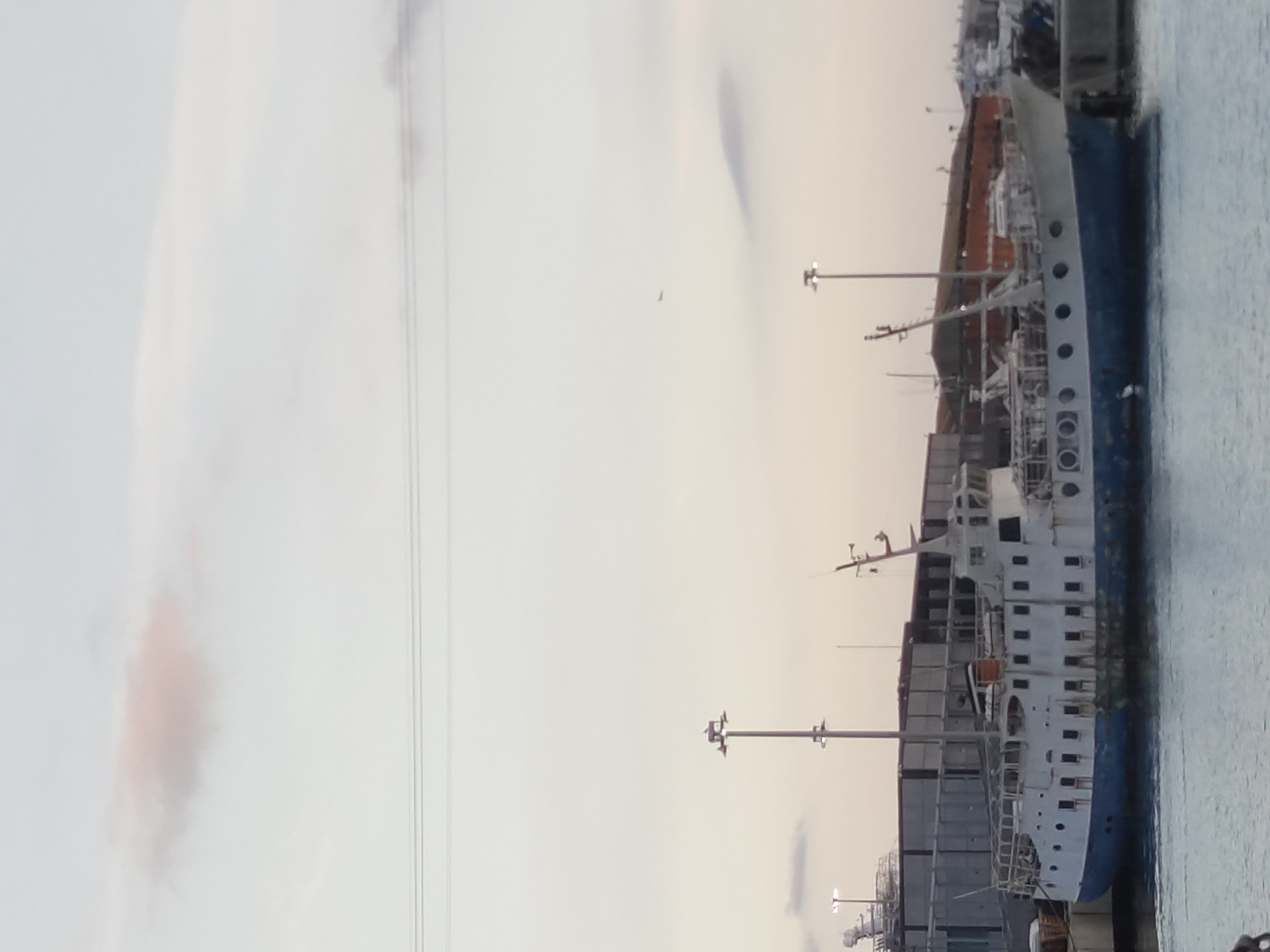 An aging, white and blue passenger ferry with multiple decks and rows of windows is docked at a harbor, likely in Barcelona, Spain. The ferry appears weathered, with some rust and visible damage. In the background, industrial buildings, possibly warehouses or shipping containers, line the waterfront. Tall light poles stand near the ferry. The sky is overcast with a hint of pinkish clouds, suggesting either dawn or dusk. The water is calm. No people are clearly visible in the scene.