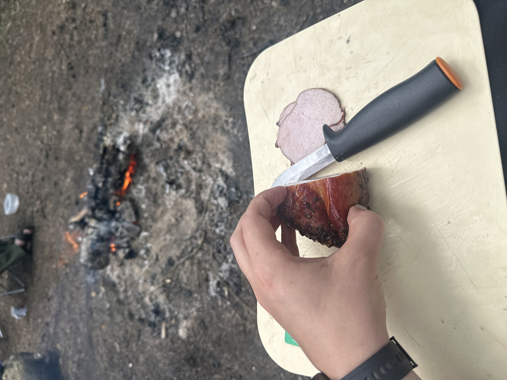 The image shows a person slicing a piece of cooked meat outdoors. The person is holding a chunk of meat with a browned exterior in their hand. A knife with a black handle and orange butt cap is being used to slice the meat. Sliced pieces are visible on a light-colored cutting board. In the background, there is a campfire burning in a dirt and ash area. There's also what looks like a camping chair nearby. The scene appears to be taking place outdoors, possibly in a wooded area near the city of Юзуфоўскі сельскі Савет, Belarus, due to the presence of a campfire and outdoor setting.