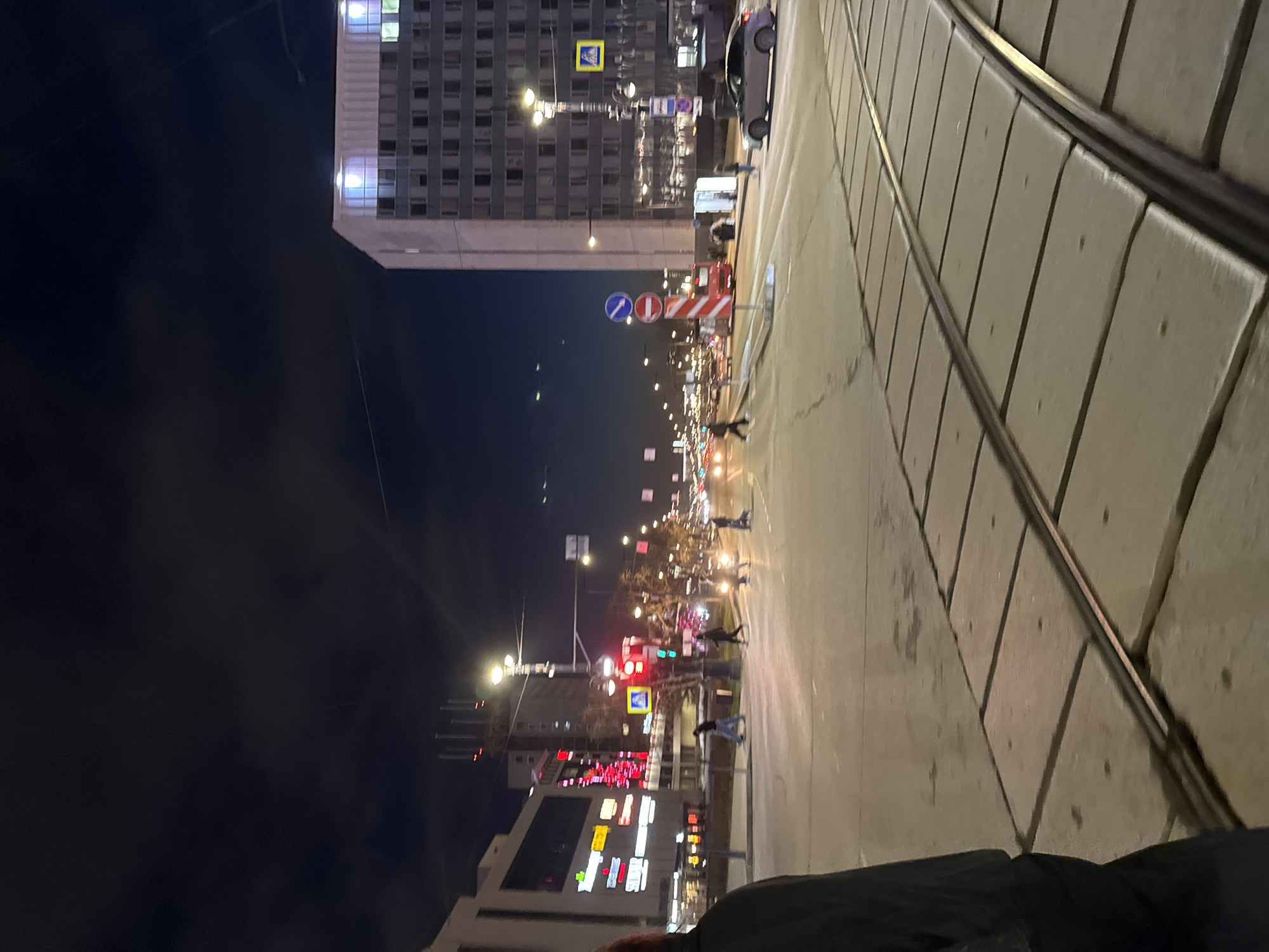 The image depicts a nighttime urban scene in Perm, Russia. In the foreground, tram tracks are visible on a concrete walkway. A person wearing a dark jacket is partially visible in the bottom right.
In the middle ground, a busy street with multiple lanes and pedestrian crossings is bustling with activity. Several people are crossing the street at a crosswalk. Numerous cars and streetlights illuminate the scene. Traffic lights are red.
In the background, buildings line the street. A tall, modern building with many windows stands out, along with another building that features bright, illuminated signs. The sky is dark, and the overall atmosphere suggests a lively city center at night. A car is parked near the curb. Street signs and traffic control devices are also present.
