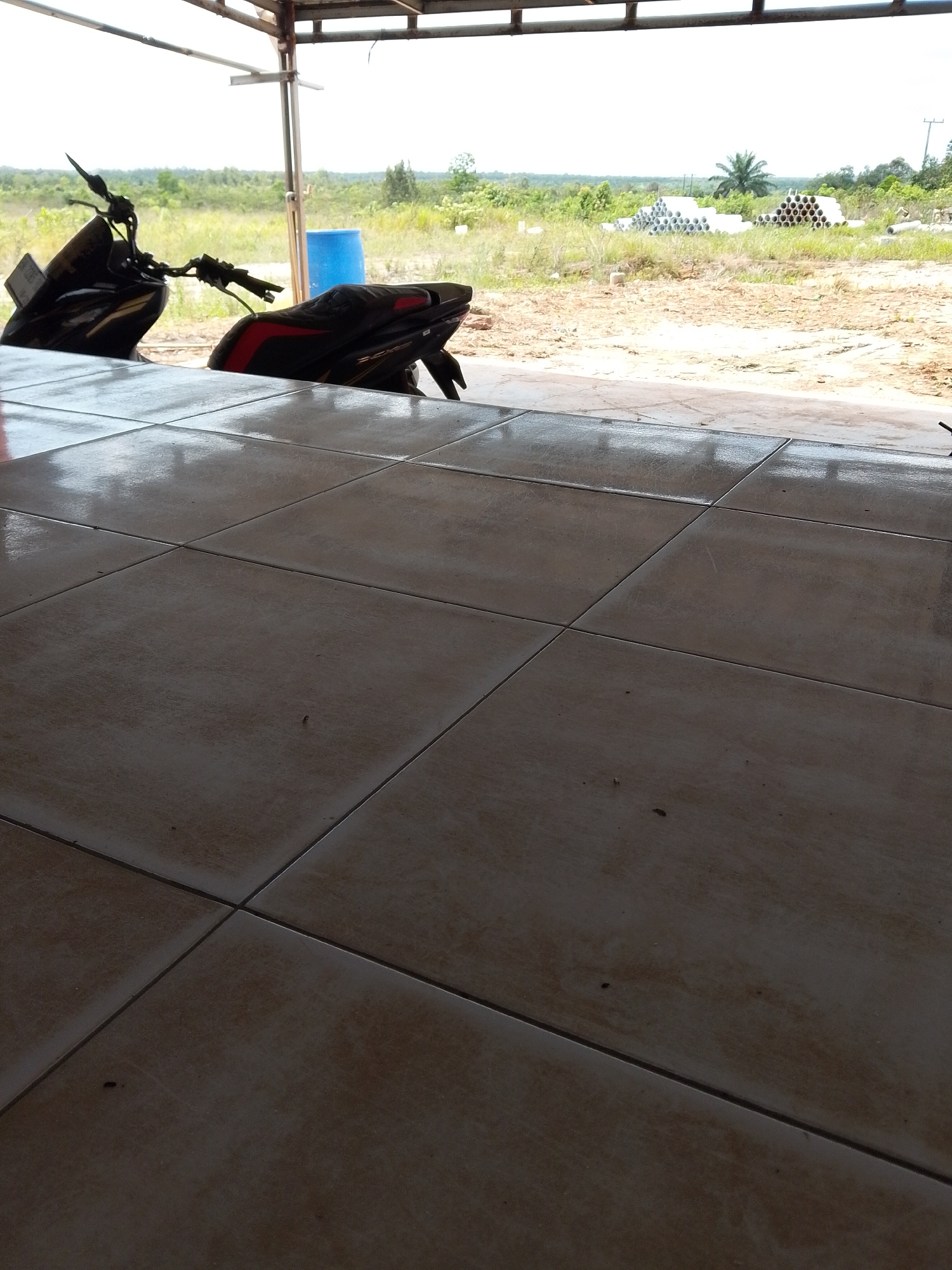 The image depicts a motorcycle parked under a shelter in Bukit Ketok, Indonesia. The foreground consists of tiled flooring, transitioning into a view of an outdoor area. The motorcycle is partially visible, with the front end and seat facing away from the viewer. Beyond the shelter, there is a grassy field with stacks of cylindrical objects (possibly pipes or construction materials) in the distance. A blue container is also visible near the motorcycle. The sky is overcast, providing ample natural lighting.