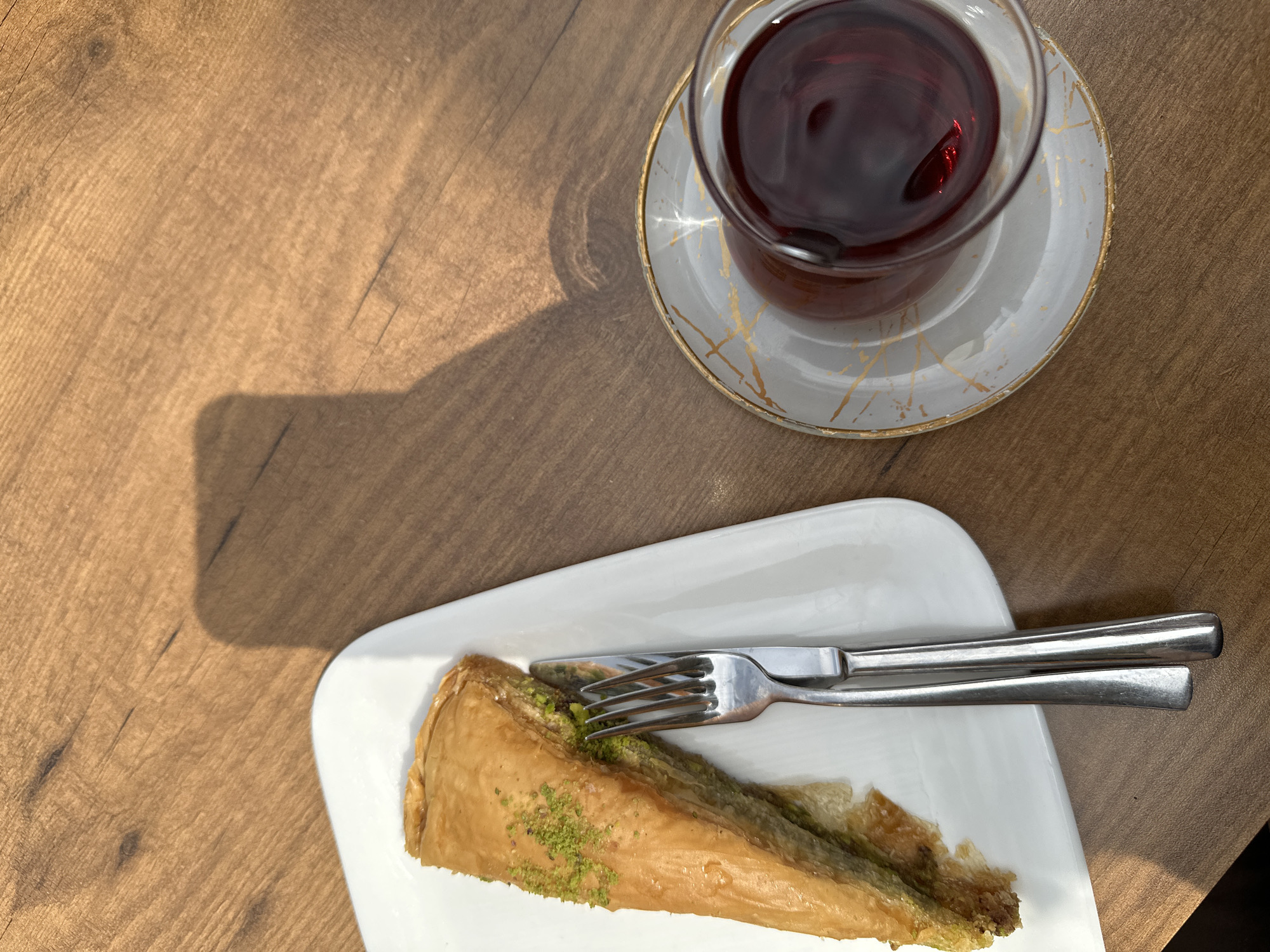 An overhead shot captures a sweet treat and drink arranged on a light brown wooden table. A triangular slice of baklava, characterized by its golden-brown, flaky layers and a generous topping of finely crushed green pistachios, is presented on a rectangular white plate. A silver-colored fork and knife are neatly placed on the plate alongside the dessert.
In the upper right portion of the frame, a clear, handleless glass cup, typical for serving Turkish tea, holds a dark red beverage. The cup rests on a white saucer featuring subtle gold, marbled patterns.
Strong, elongated shadows are cast across the left side of the wooden table, suggesting bright daylight, indicating the photo was taken on a sunny morning or afternoon. The table surface displays visible wood grain. The setup suggests a relaxing pause for a snack or dessert, consistent with a cafe or restaurant setting in Fethiye, Turkey. No text is visible in the image.